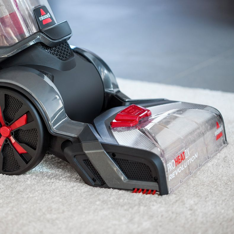 Zbliżenie odkurzacza Bissell ProHeat 2X Revolution na białym dywanie. Odkurzacz jest szary i czarny z czerwonymi akcentami.