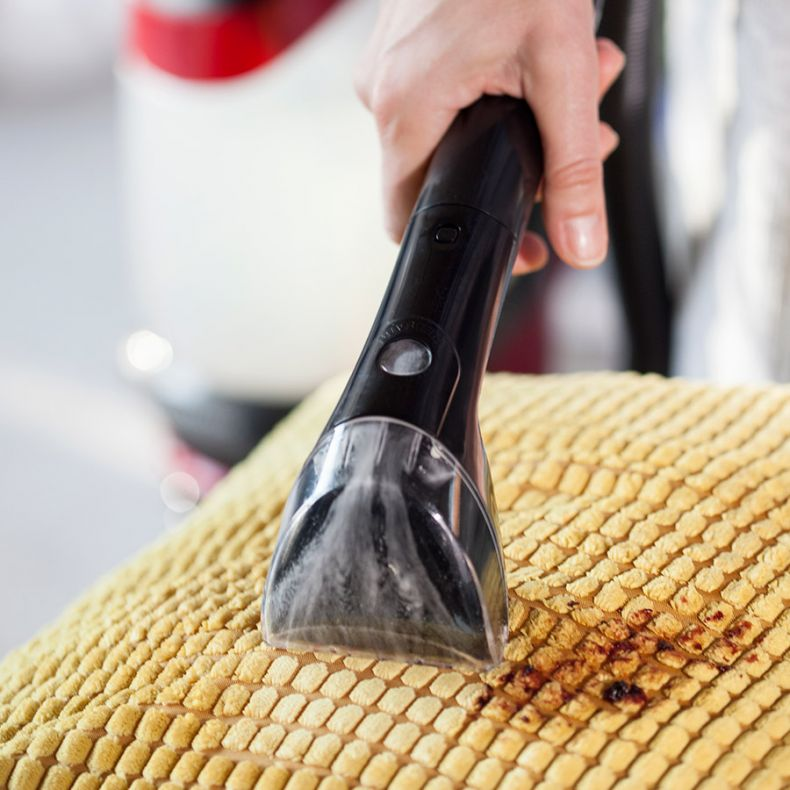 Ręka trzyma czarny odkurzacz do tapicerki nad żółtą tkaniną z brązową plamą, czyszcząc tkaninę.