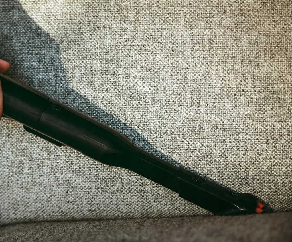 Czarna głowica odkurzacza czyści szarą kanapę z tkaniny. Odkurzacz jest pod kątem, w użyciu. Tłem jest faktura kanapy.