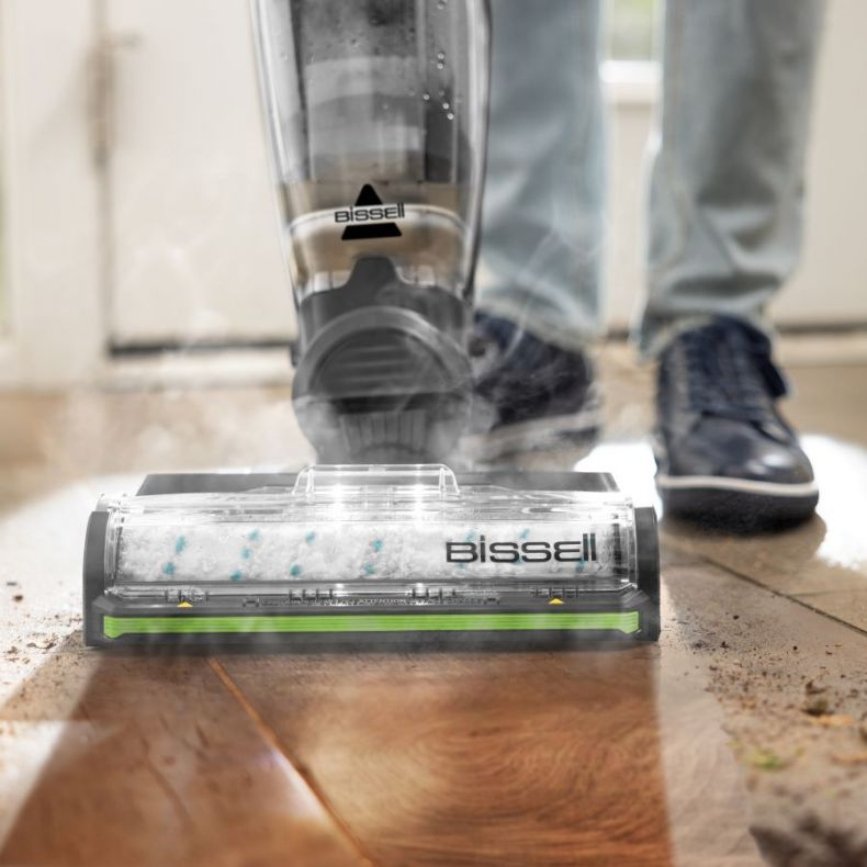 Osoba używa odkurzacza Bissell na drewnianej podłodze. Odkurzacz jest czarny i przezroczysty. Widać parę. Osoba ma na sobie dżinsy i buty.