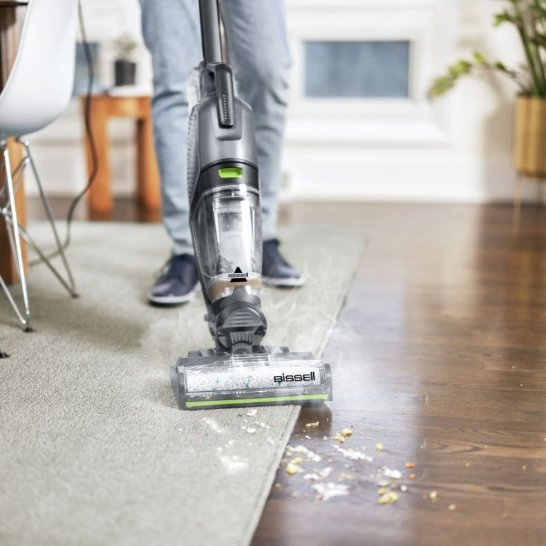 Osoba odkurza dywan odkurzaczem Bissell. Na dywanie i podłodze z twardego drewna widać zanieczyszczenia. Odkurzacz jest czarny i szary.