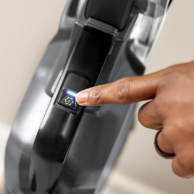 Palec naciska przycisk na szarym odkurzaczu. Przycisk świeci niebieskim światłem. Na palcu jest czarny pierścień.