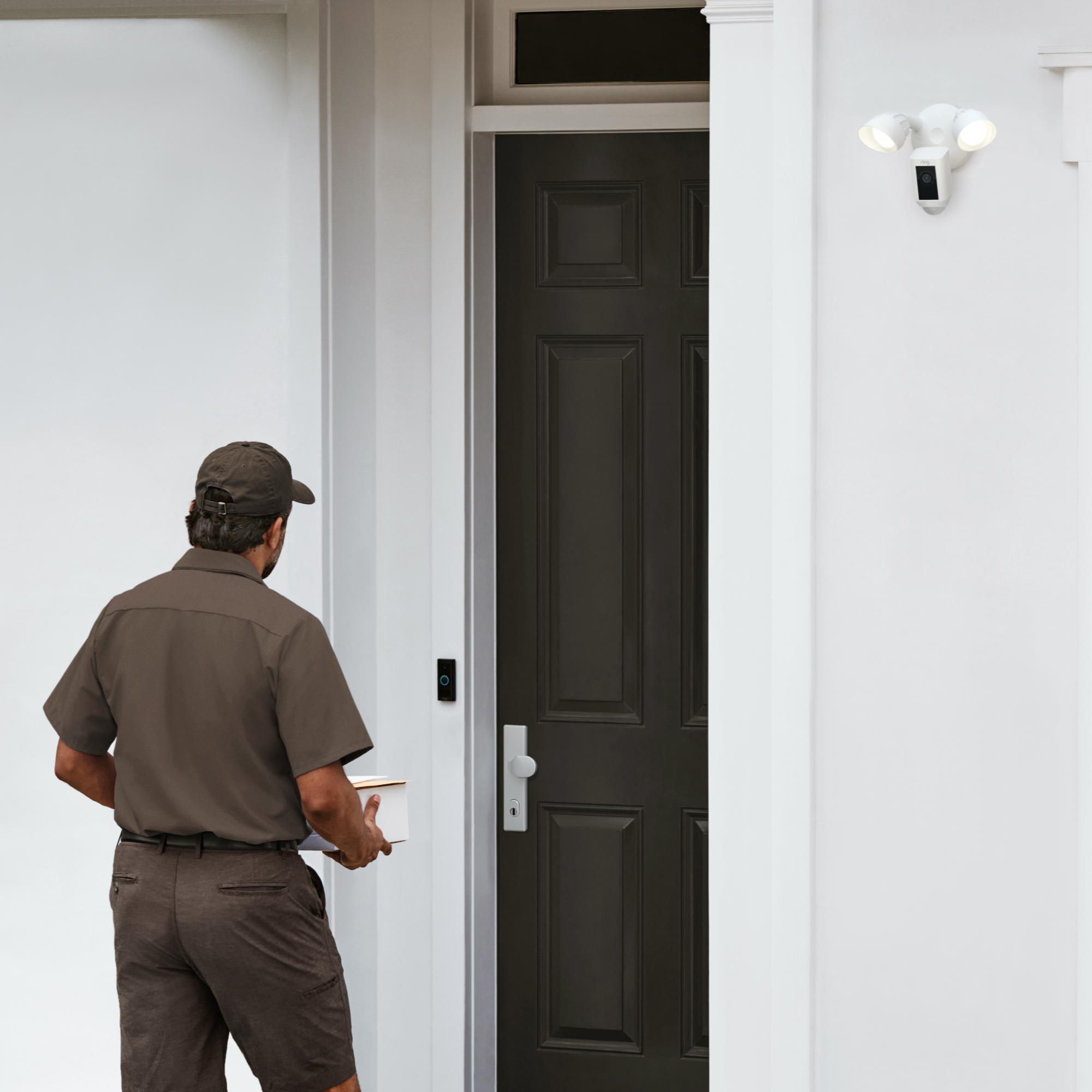 Zusteller in brauner Uniform nähert sich einer dunklen Tür. Über der Tür ist eine Überwachungskamera montiert.