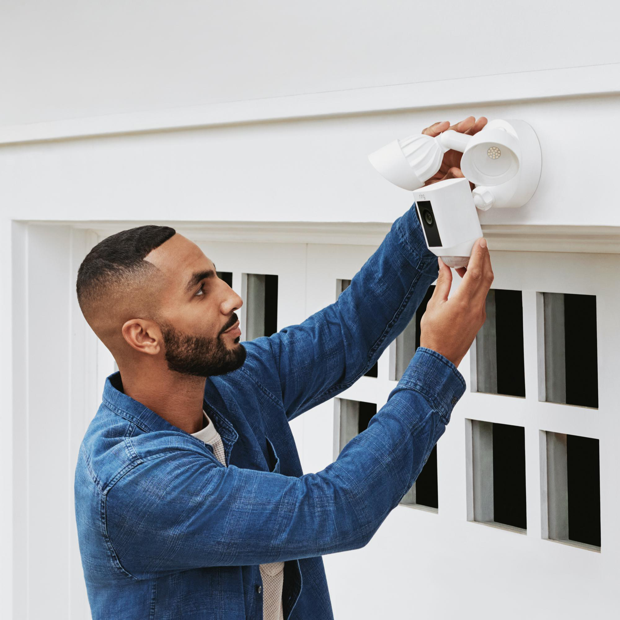 Ein Mann im blauen Hemd installiert eine weiße Überwachungskamera an einer weißen Wand. Die Kamera befindet sich in der Nähe eines Fensters.
