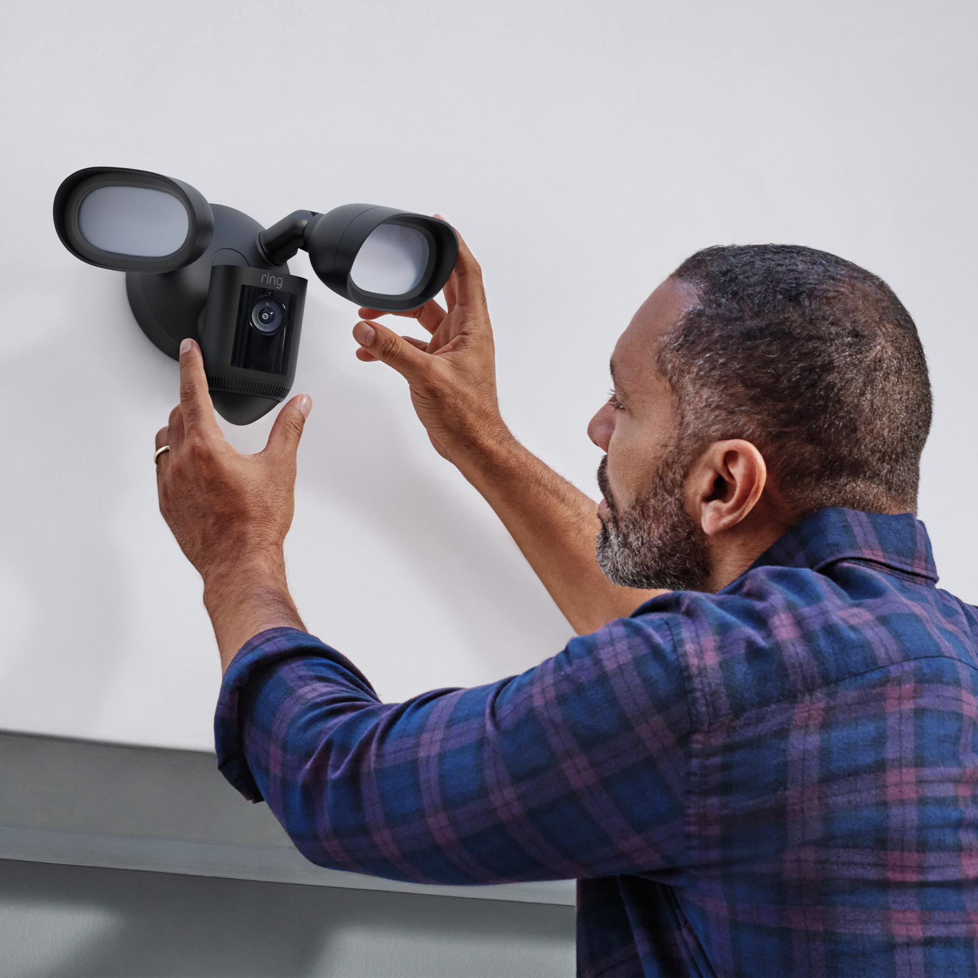 Ein Mann installiert eine schwarze Sicherheitskamera mit Lichtern an einer weißen Wand. Er trägt ein kariertes Hemd.