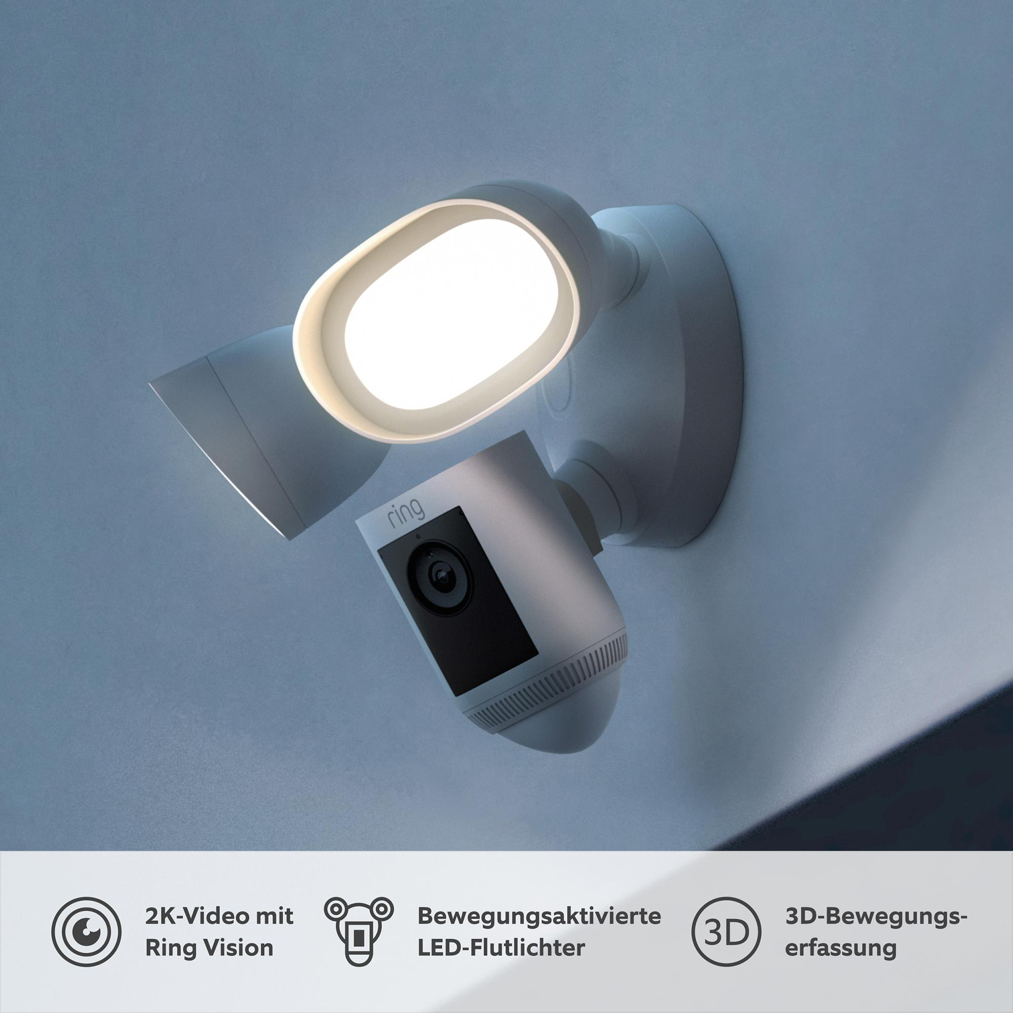 Eine weiße Ring-Flutlichterkamera an einer Wand. Sie hat zwei Lichter und eine Kamera. Text auf dem Bild.