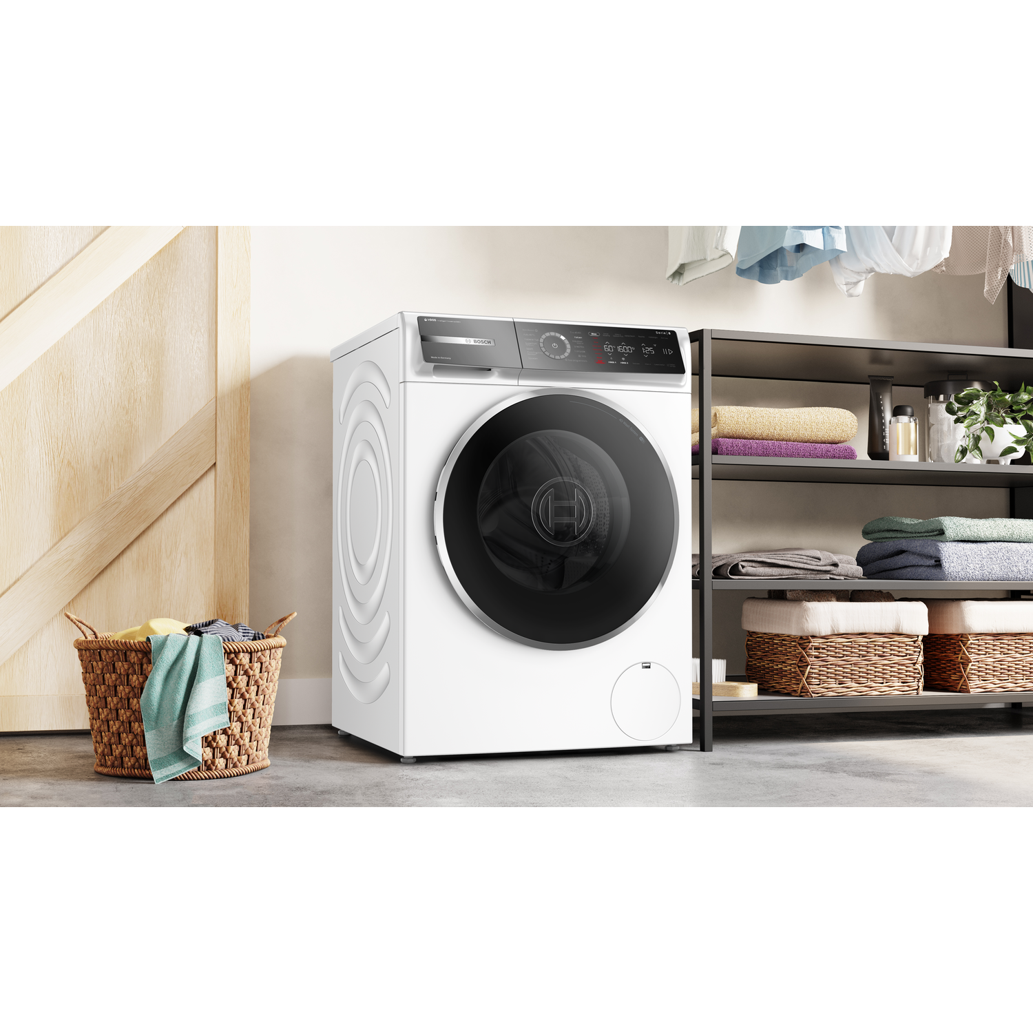 A white washing machine stands next to a wooden door and a metal shelf with towels and baskets. A basket with towels is on the floor.