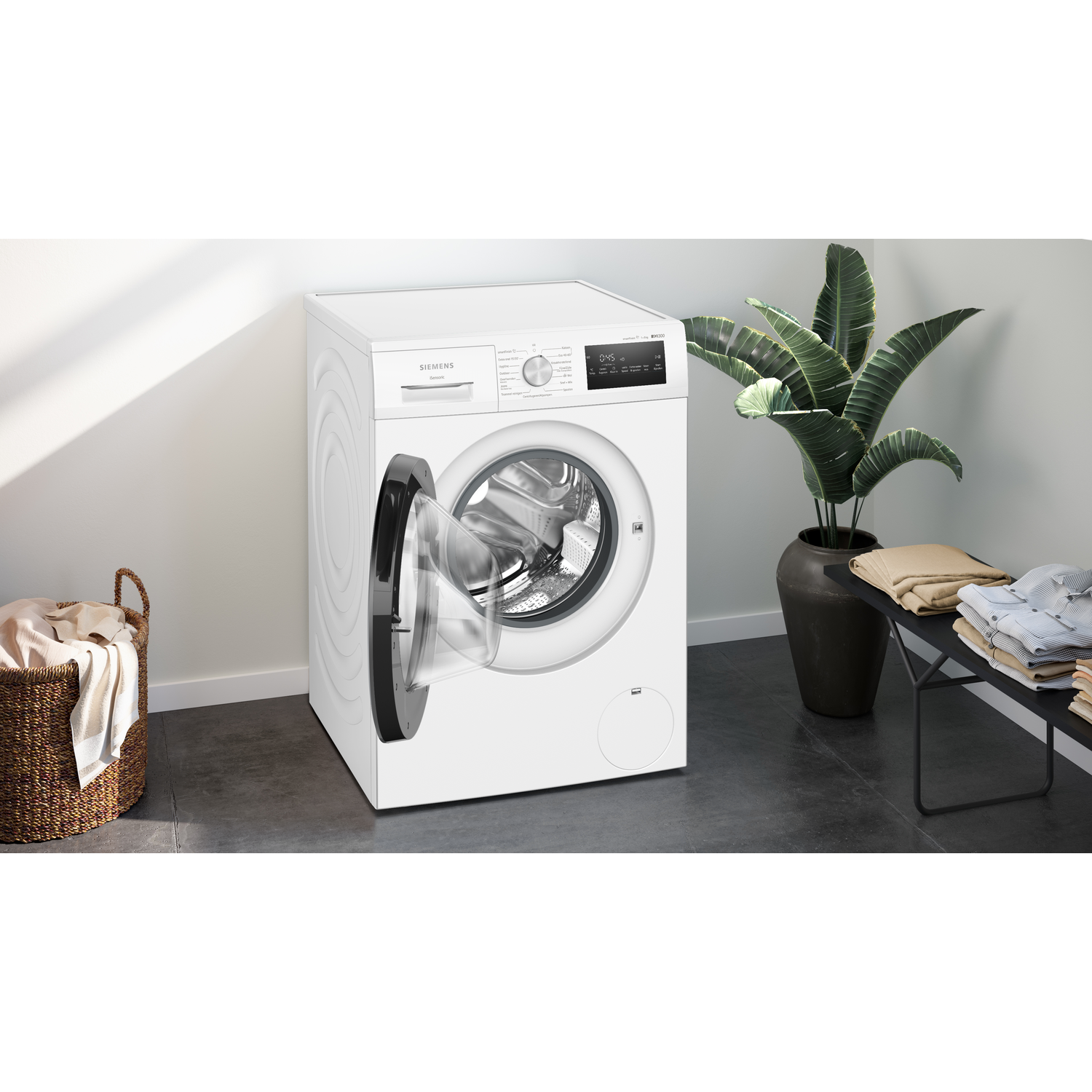 A white washing machine with an open door. A basket of clothes on the left. A plant and folded clothes on the right.