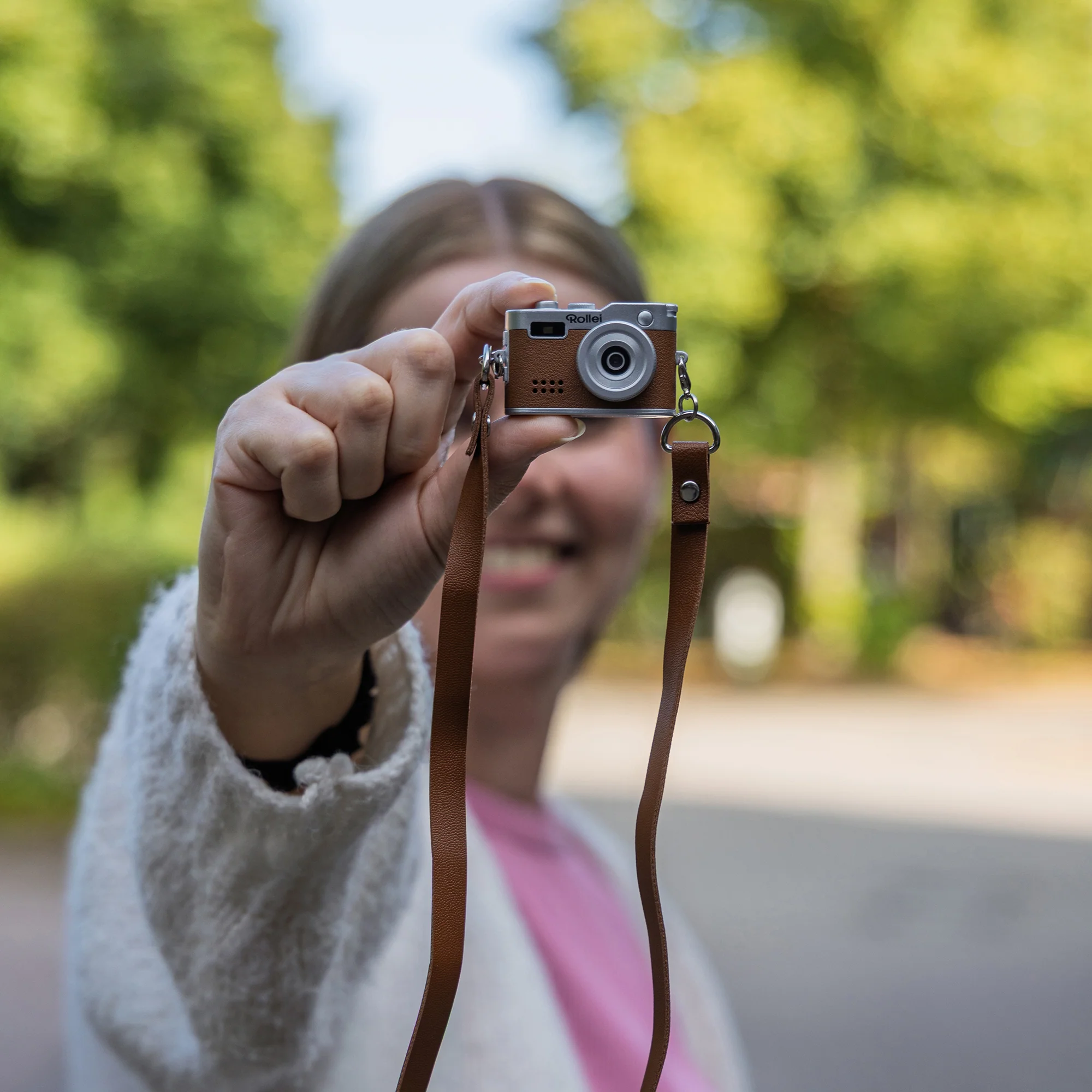 Eine Frau hält eine Miniaturkamera. Die Kamera ist braun und silber, mit einem braunen Riemen. Bäume sind im Hintergrund.