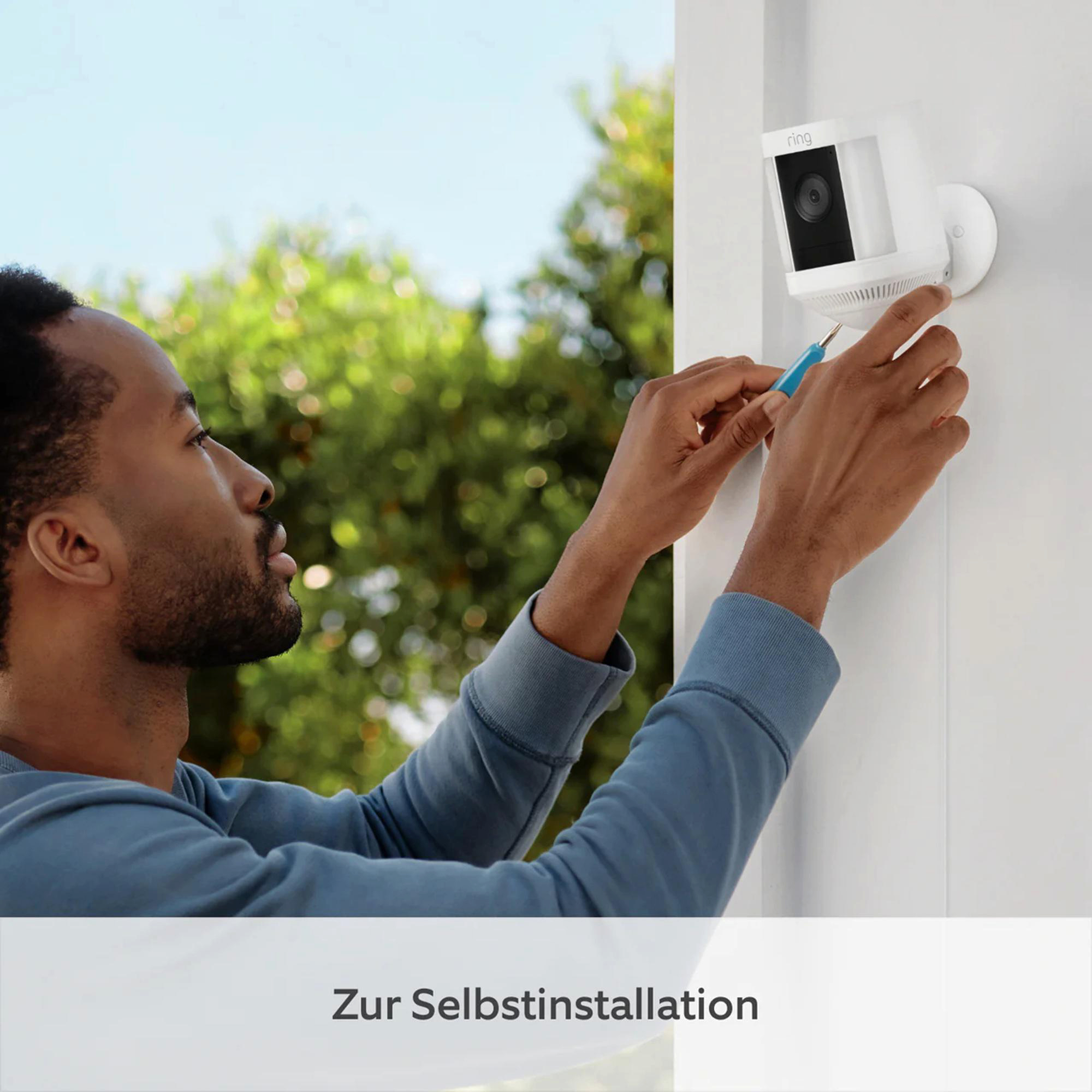 Ein Mann installiert eine weiße Überwachungskamera an einer Wand. Er hält einen Schraubendreher. Der Hintergrund zeigt grüne Bäume und blauen Himmel.
