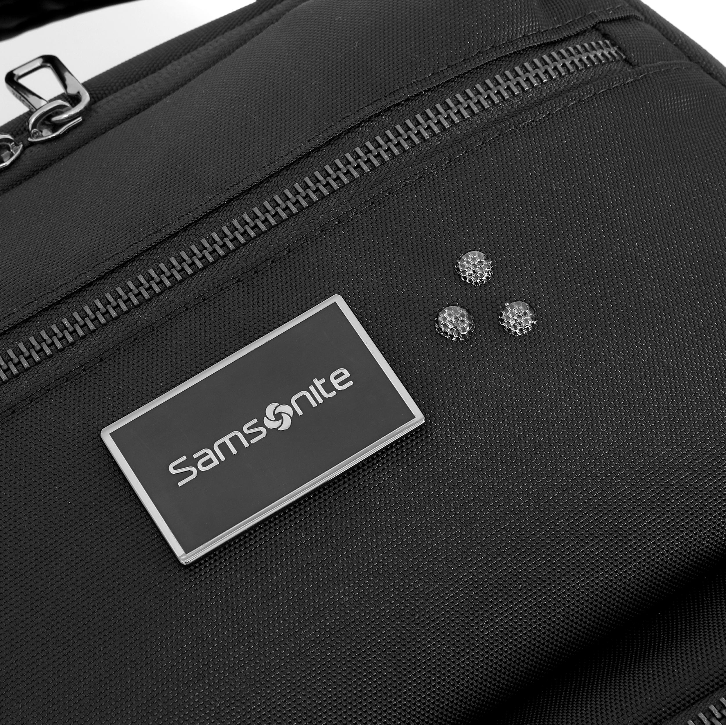 Siyah bir Samsonite çantasının yakın çekimi. Fermuar, logo ve dekoratif çiviler içerir. Çanta dokulu bir yüzeye sahiptir.