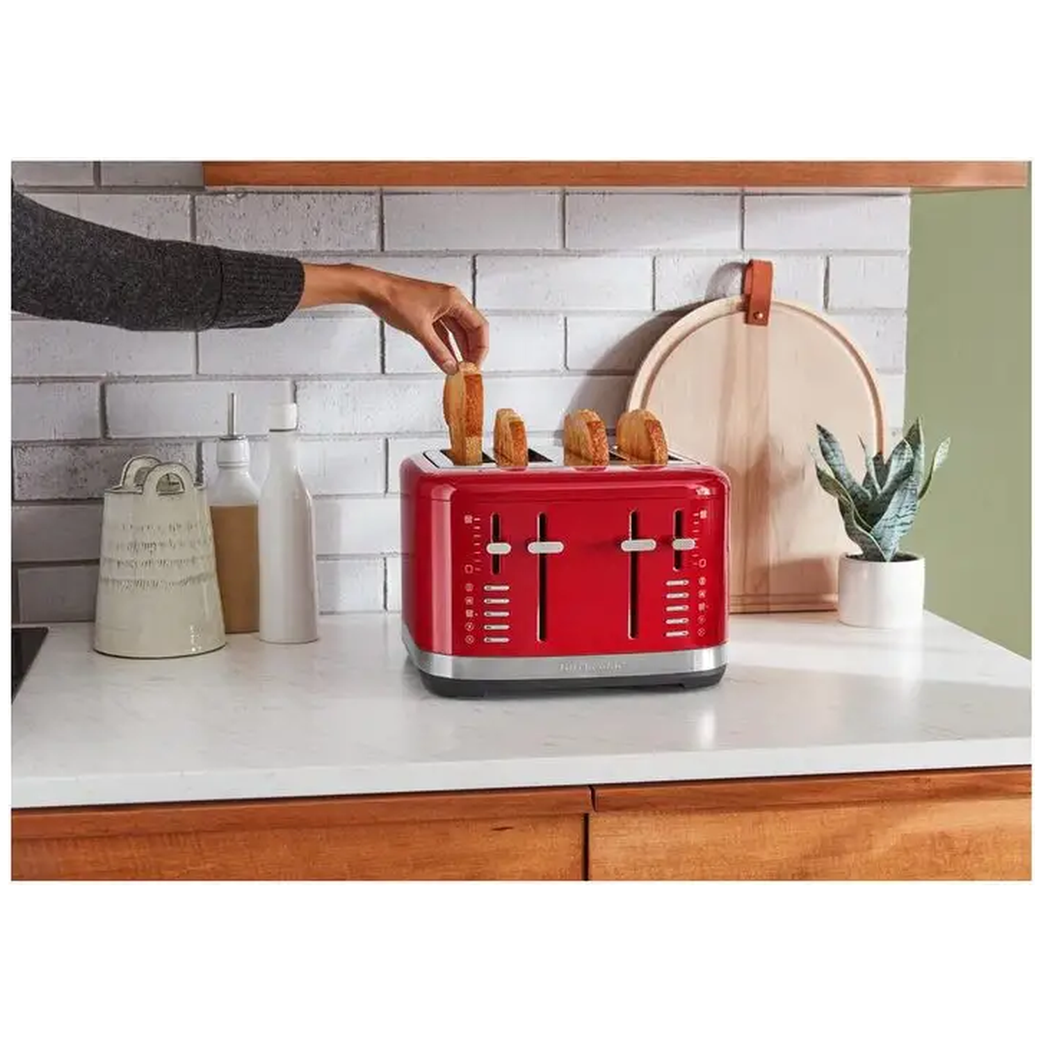 Een hand haalt toast uit een rode broodrooster. Andere elementen: snijplank, plant, witte flessen en een bakstenen muur.