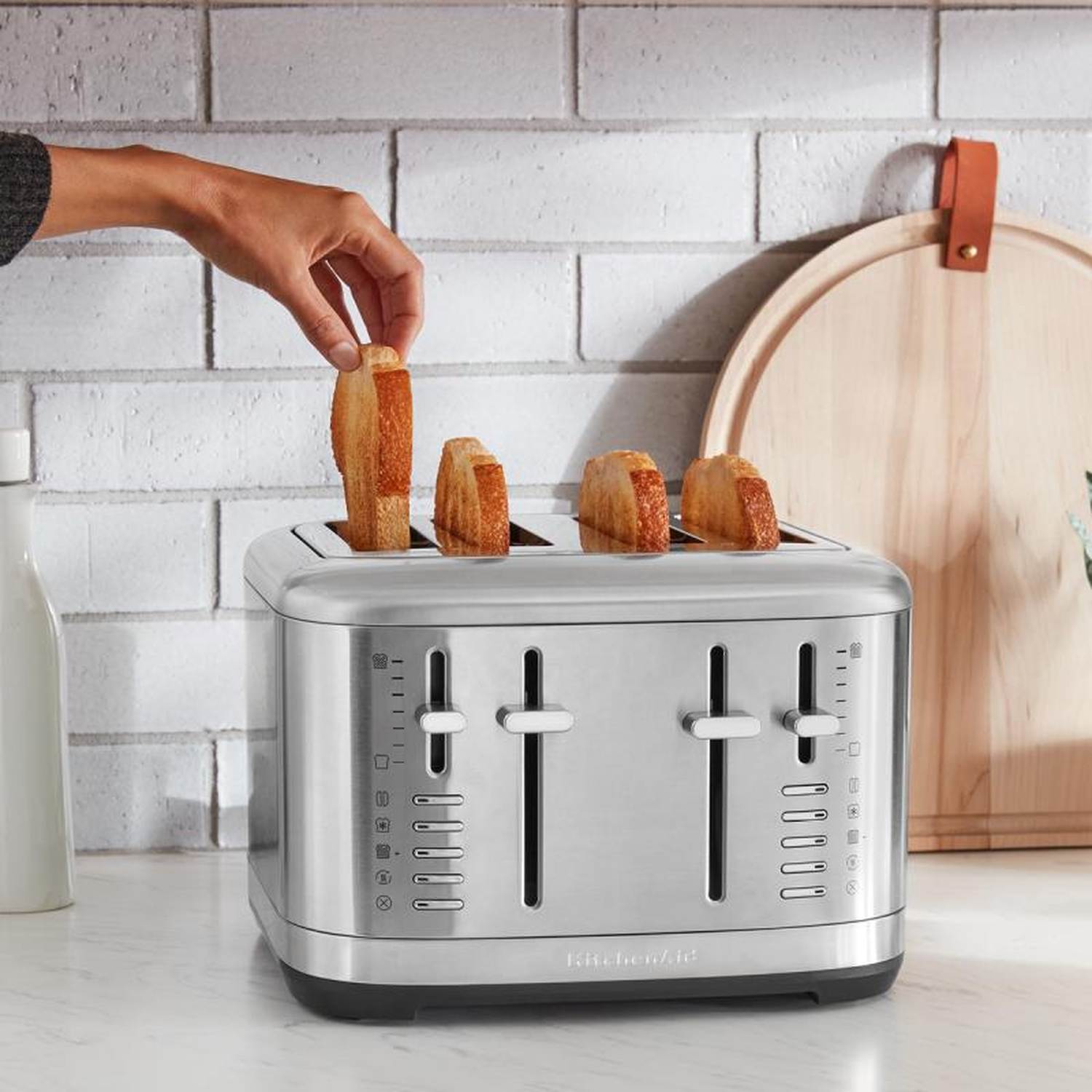 Een hand plaatst toast in een zilveren broodrooster. Drie sneetjes toast zitten al in de broodrooster. Een houten snijplank hangt op de achtergrond.