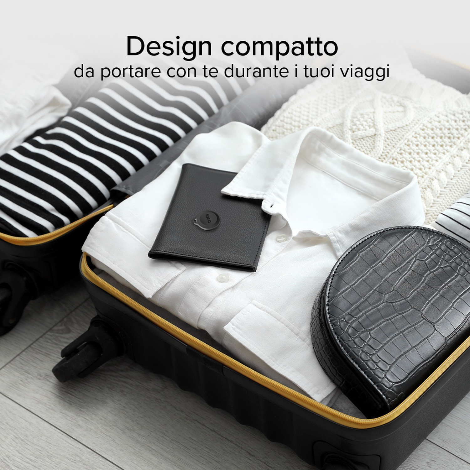 Valigia piena di vestiti, un portafoglio nero e un astuccio nero strutturato. La valigia è su un pavimento di legno.