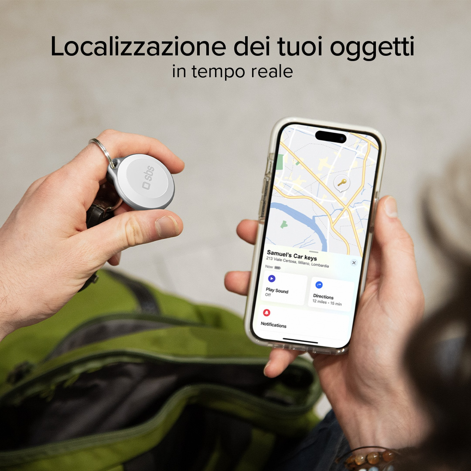 Una persona tiene in mano un telefono che mostra una mappa con la posizione delle chiavi dell'auto. Un localizzatore bianco è attaccato alle chiavi. Uno zaino verde è sullo sfondo.