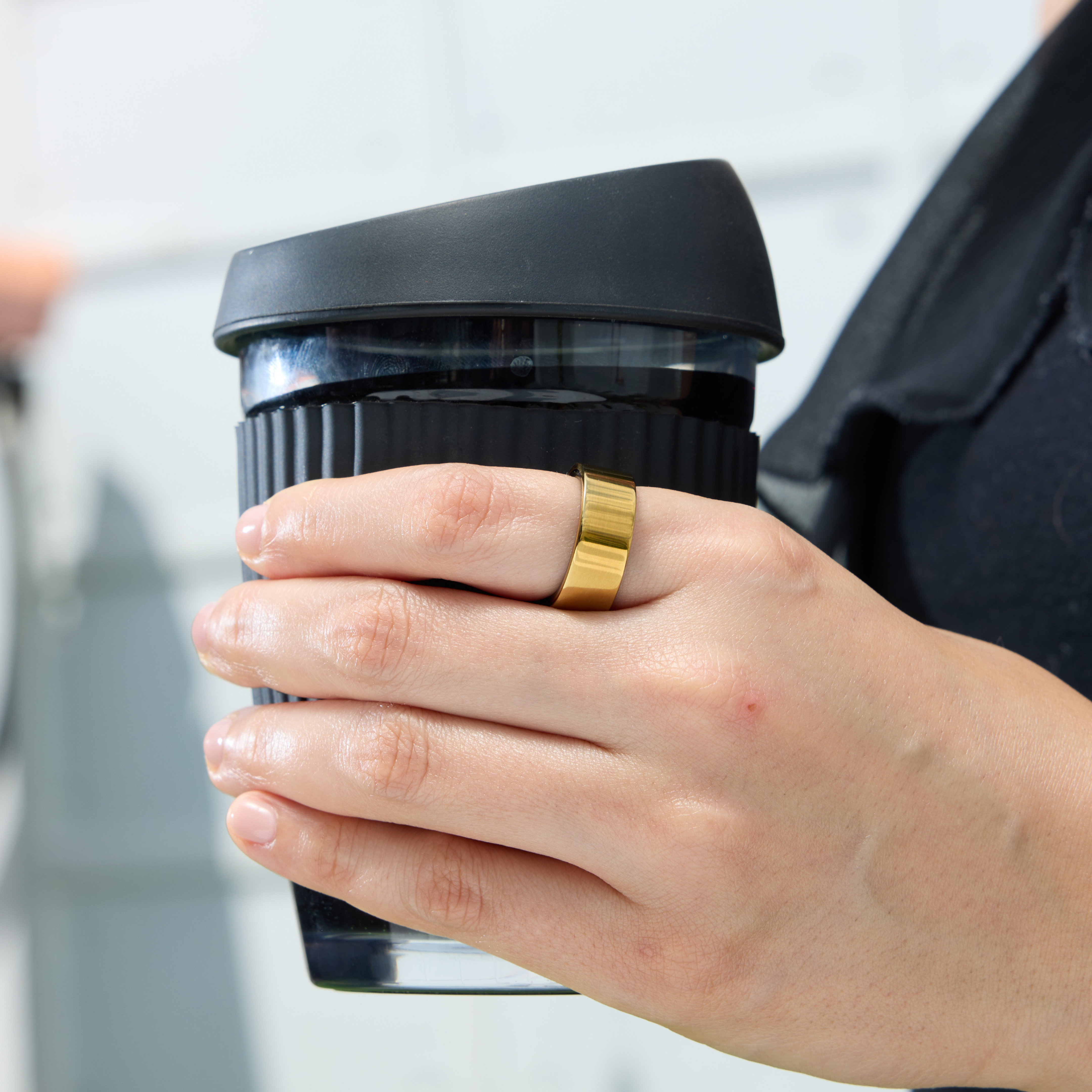 Eine Person hält einen schwarzen Reisebecher. Ein goldener Ring befindet sich an einem Finger. Der Hintergrund ist verschwommen.