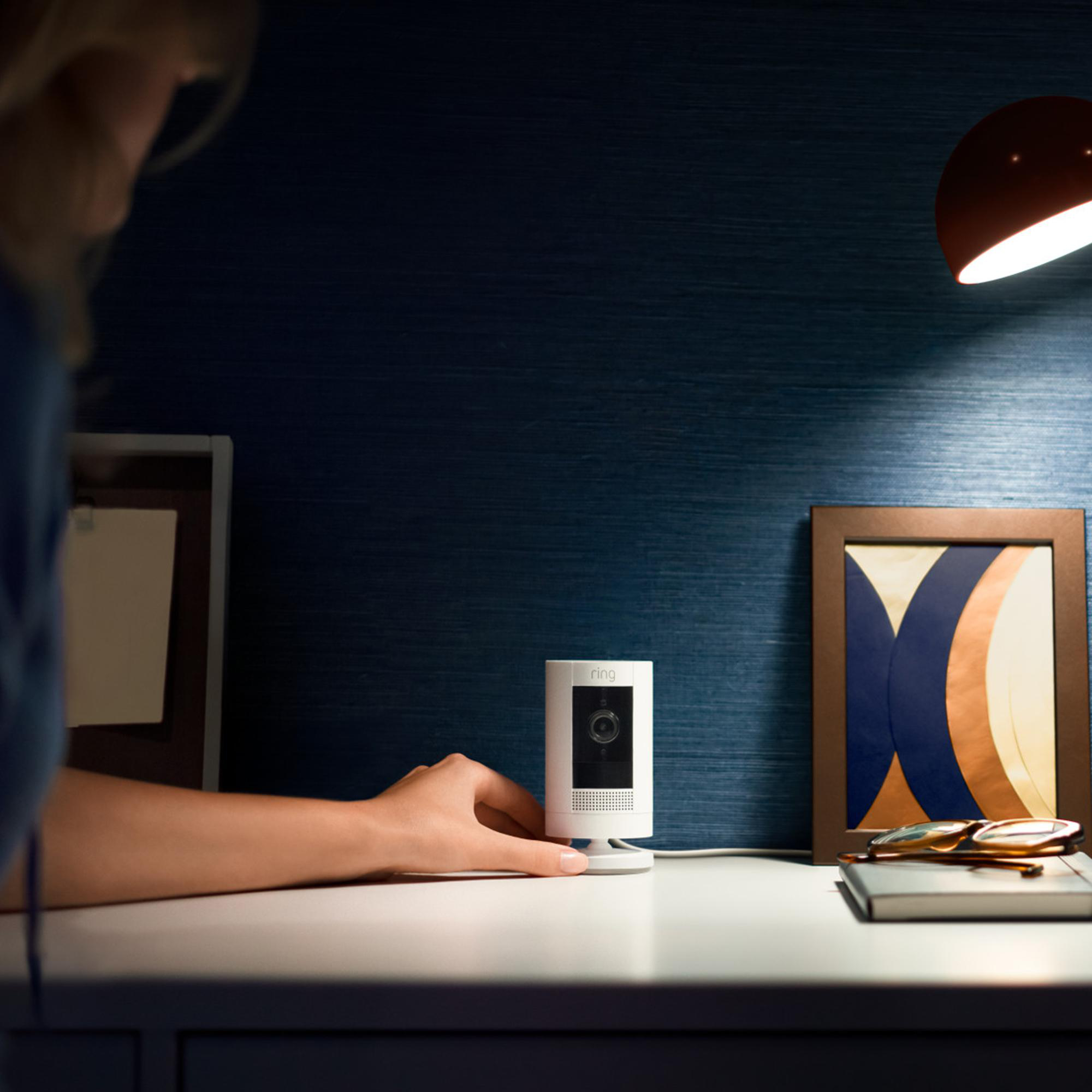 Die Hand einer Frau berührt eine weiße Ringkamera. Auf dem Schreibtisch liegen ein gerahmtes Bild und eine Brille. Eine Lampe scheint Licht.