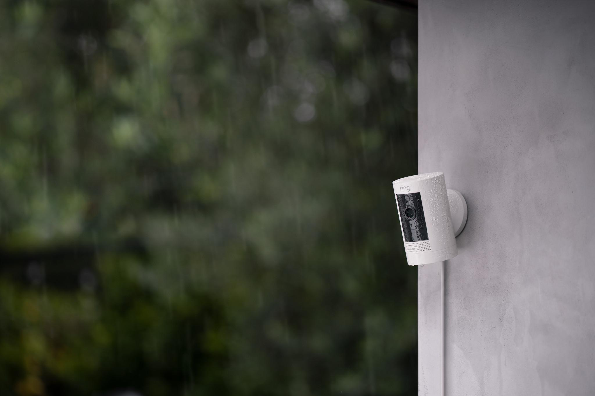 Eine weiße Ring-Sicherheitskamera ist im Regen an einer grauen Wand montiert. Im verschwommenen Hintergrund sind grüne Bäume zu sehen.