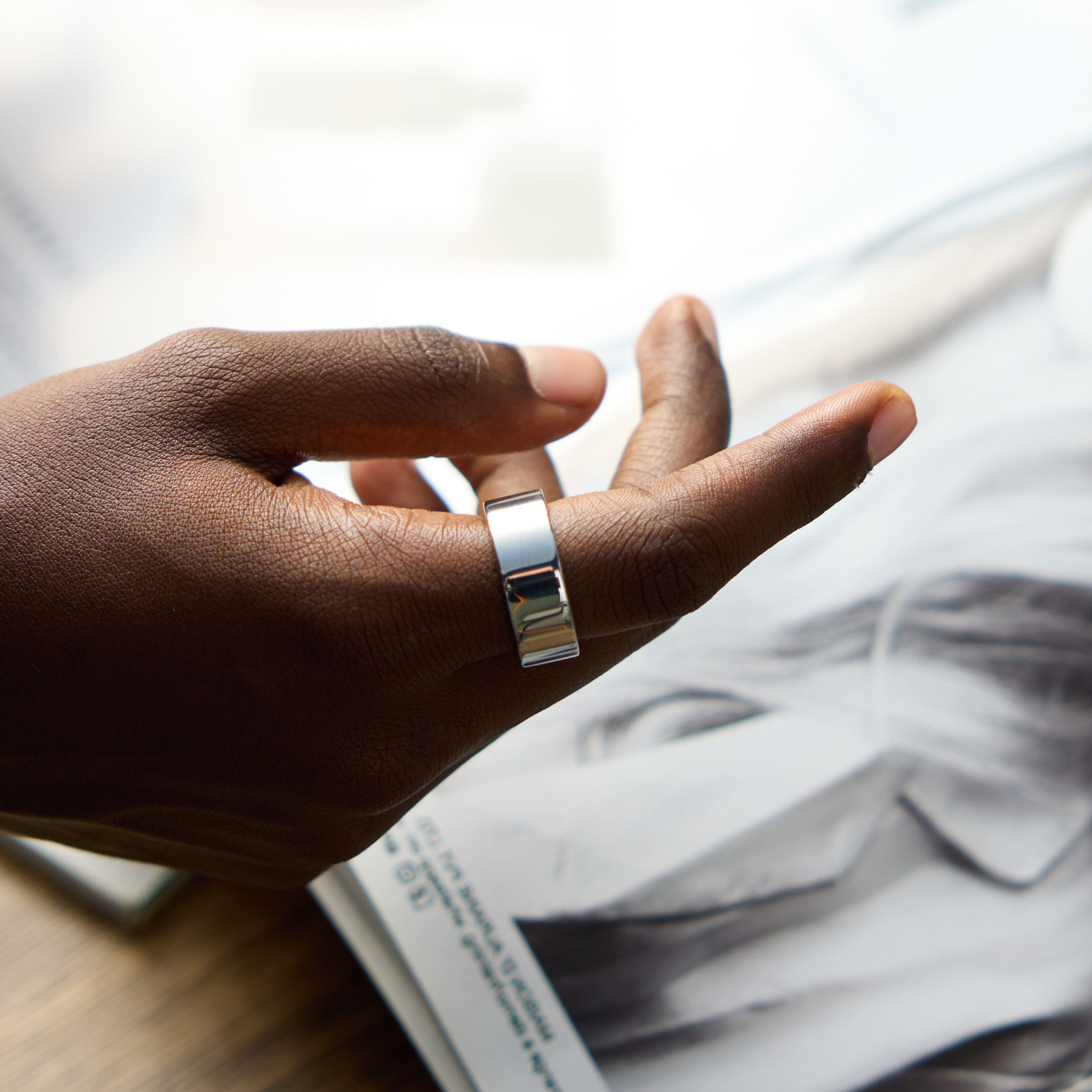 Eine Hand mit einem silbernen Ring am Ringfinger. Der Hintergrund ist ein Magazin und ein Holztisch.