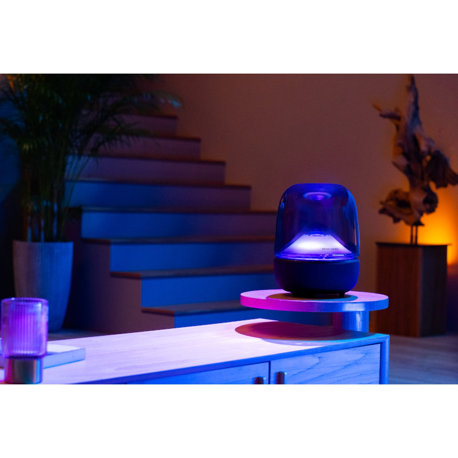 Un altoparlante blu luminoso su un tavolo di legno. Sullo sfondo, una scala e una pianta, illuminate con luci blu e viola.