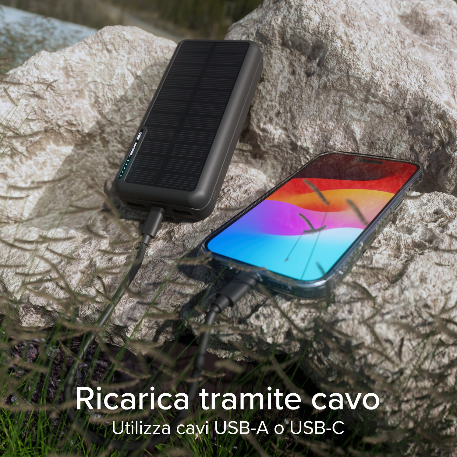 Un caricatore solare nero e uno smartphone sono collegati con cavi USB su una roccia, con erba in primo piano.