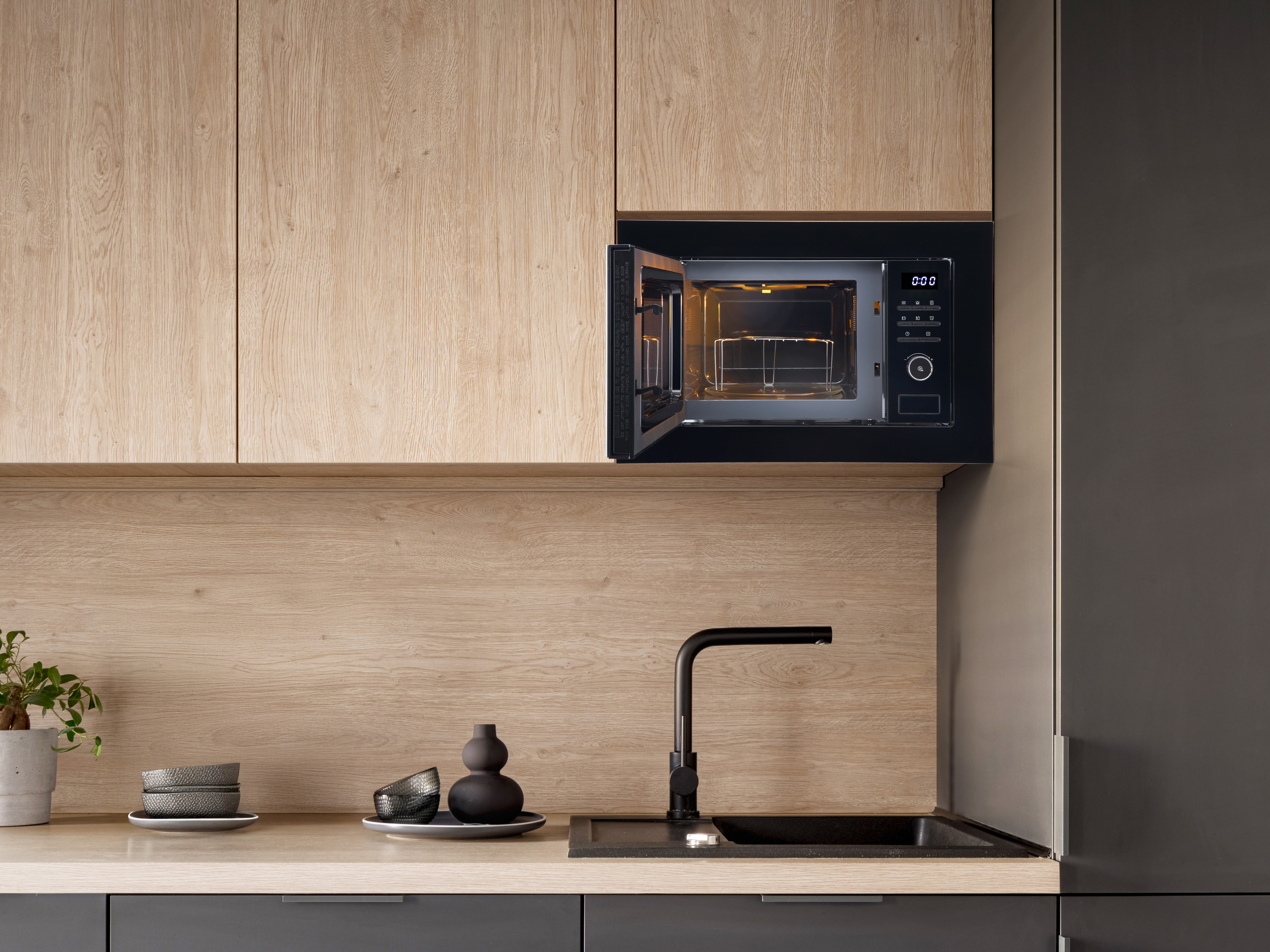Moderne Küche mit schwarzer Mikrowelle, offen. Holzschränke und Arbeitsplatte. Ein schwarzes Waschbecken und Wasserhahn.
