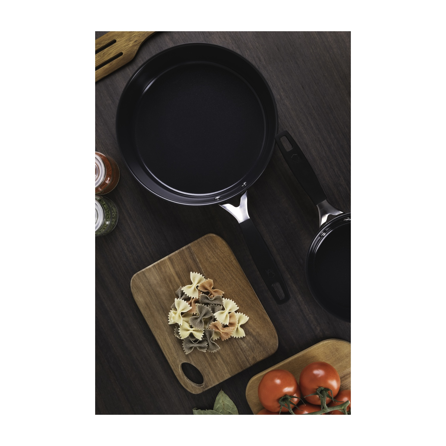 Vista dall'alto di una scena di cucina con padelle, pasta, pomodori e spezie su una superficie di legno scuro.