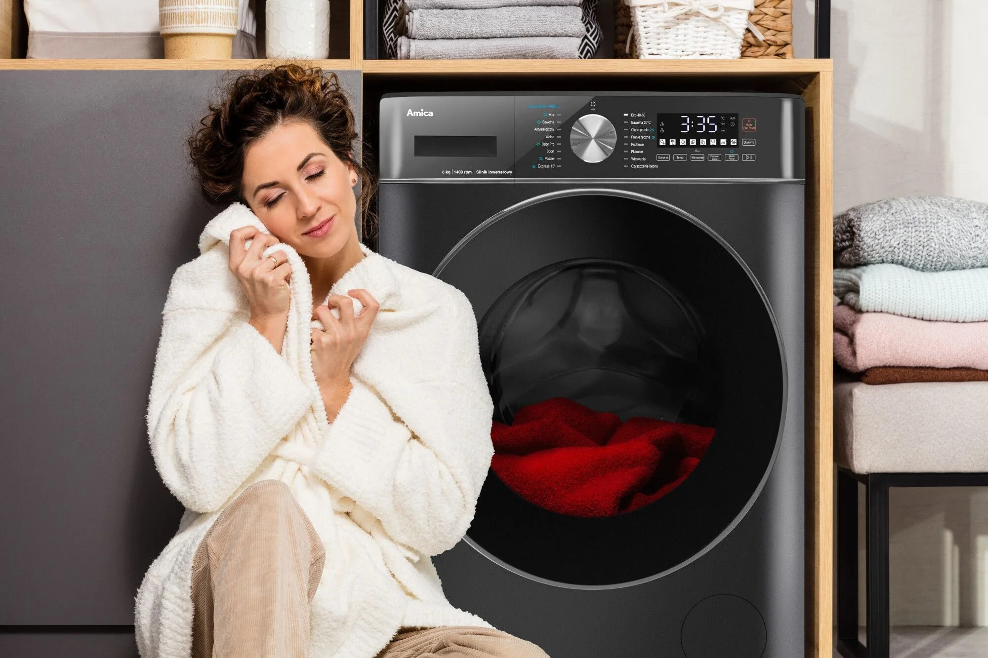 Kobieta w białym szlafroku, obok czarnej pralki z czerwonymi ubraniami. Stos ubrań po prawej.