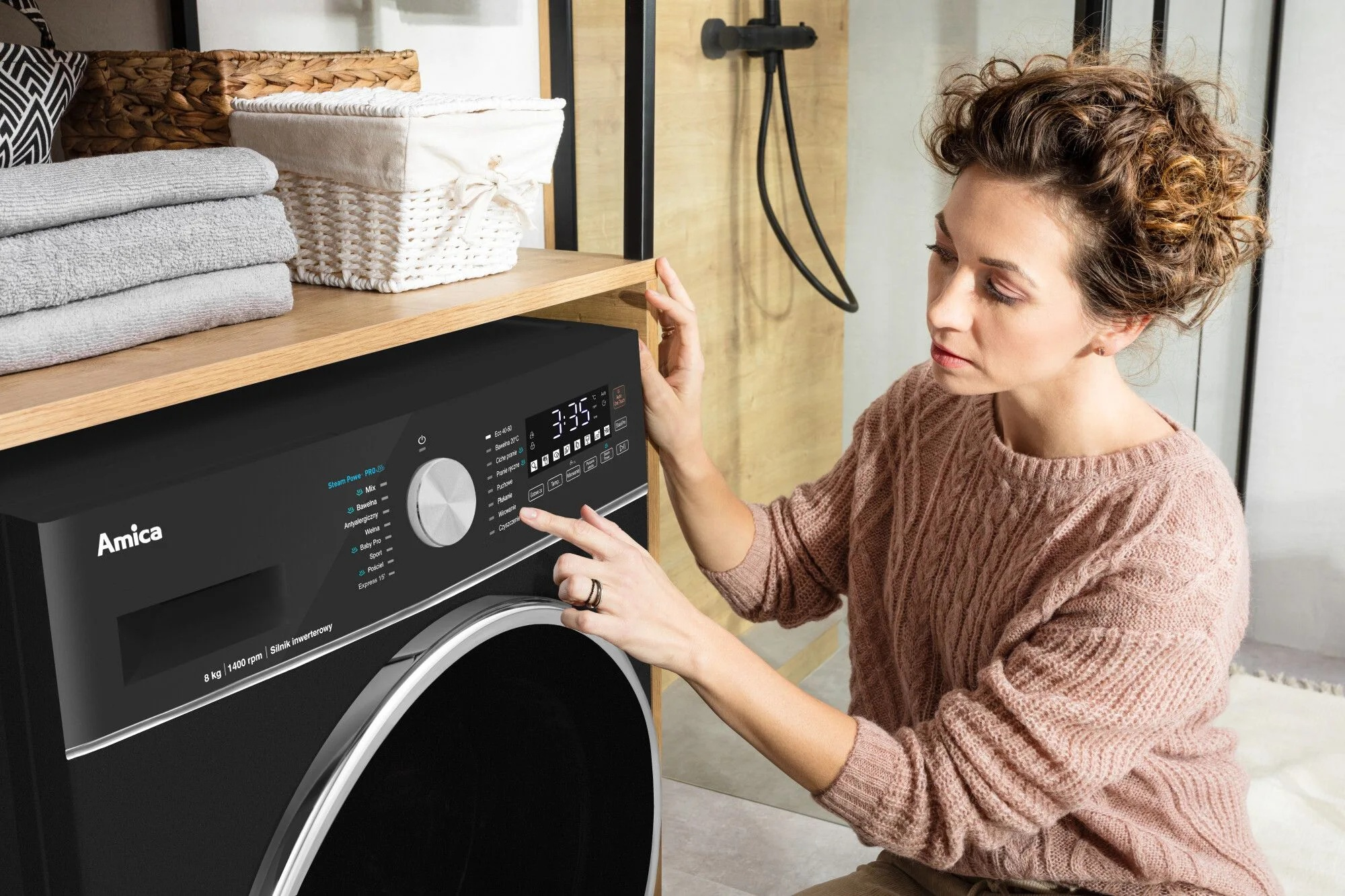 Kobieta w różowym swetrze obsługuje pralkę. Maszyna jest czarna i ma cyfrowy wyświetlacz. Ręczniki i kosze znajdują się na półce.