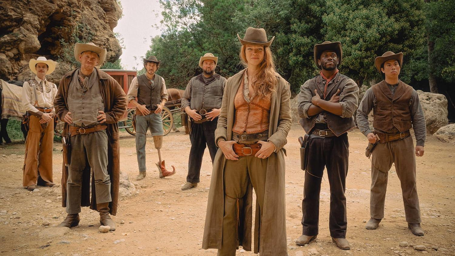 Eine Gruppe Cowboys steht im Freien. Sie tragen Hüte und Westernkleidung. Einige haben Waffen. Eine Frau steht im Zentrum.