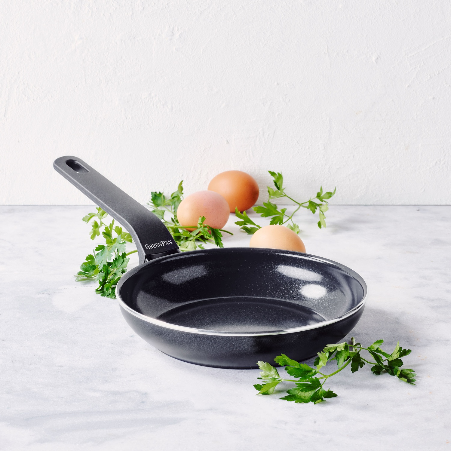 A black frying pan with a handle, eggs, and parsley on a marble surface. The pan has the brand name GREENPAN on the handle.