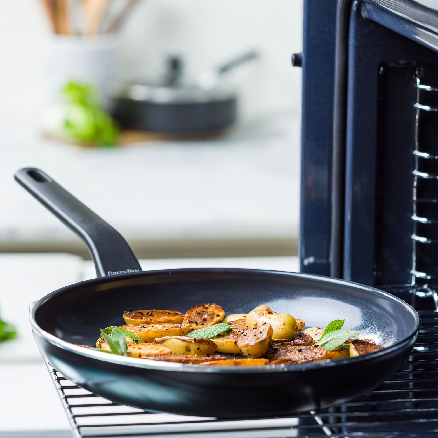 A black pan with roasted potatoes and herbs sits on a rack near an oven. Kitchen utensils are in the background.