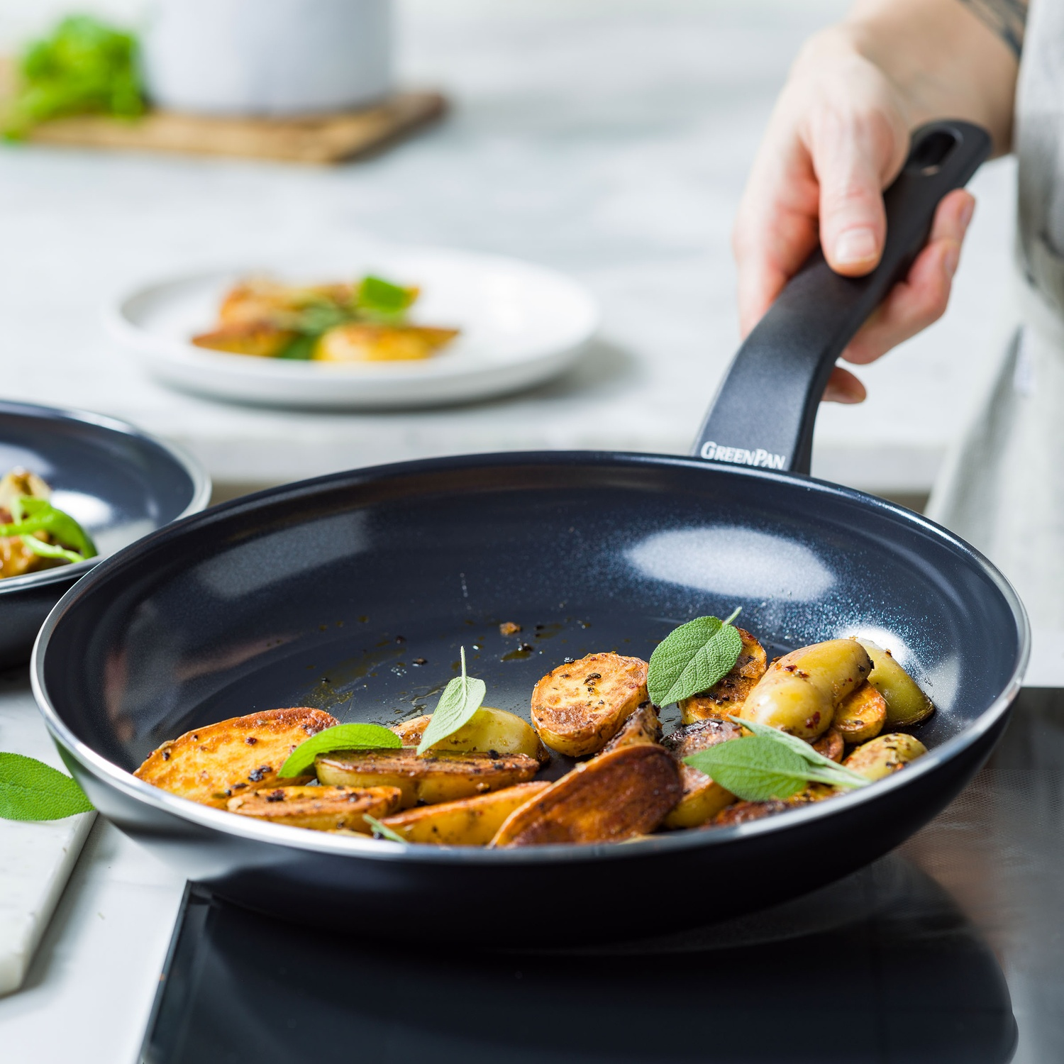 Une personne tient une poêle avec des pommes de terre frites et des feuilles de sauge. Un autre plat avec une nourriture similaire est en arrière-plan.