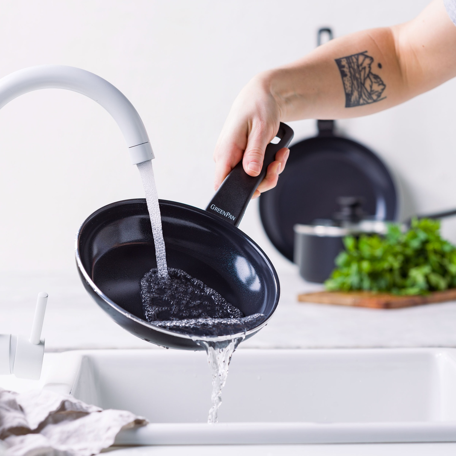 Une poêle noire est tenue sous un robinet blanc. L'eau coule dans la poêle. D'autres poêles et des herbes sont en arrière-plan.