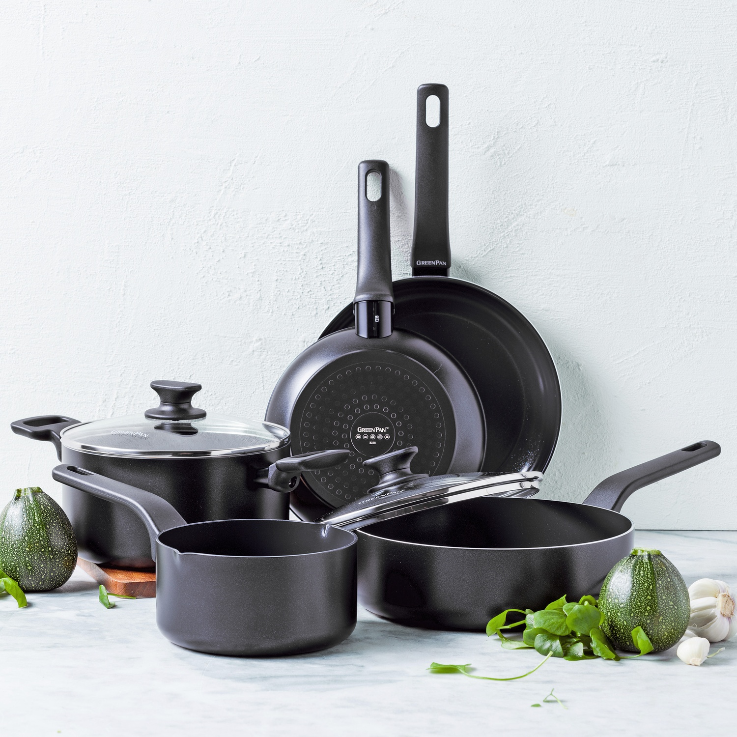 Ensemble d'ustensiles de cuisine noirs avec légumes. Poêles, casseroles et couvercles sont disposés. Courgettes, ail et herbes au premier plan.