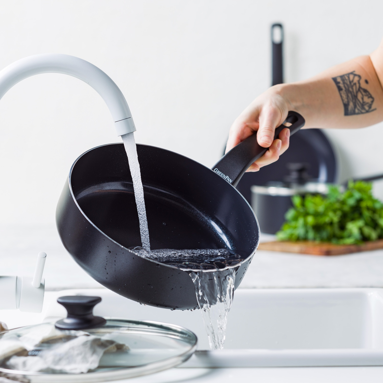 A black pan is held under a faucet, water pouring into it. A hand with a tattoo holds the pan. A sink, lid, and greens are visible.