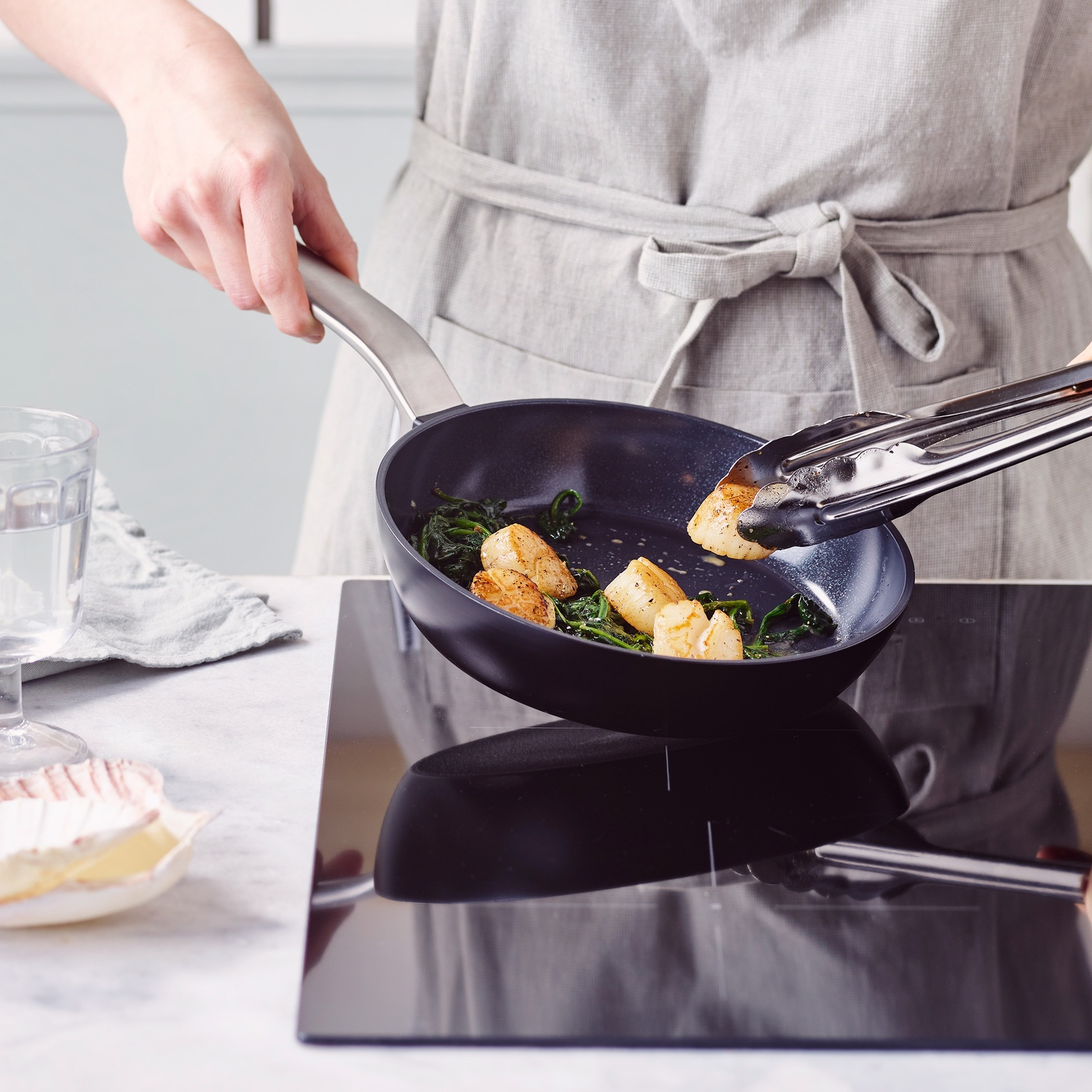 A person is cooking scallops and spinach in a black pan on a cooktop. Using tongs, the person is flipping the scallops.
