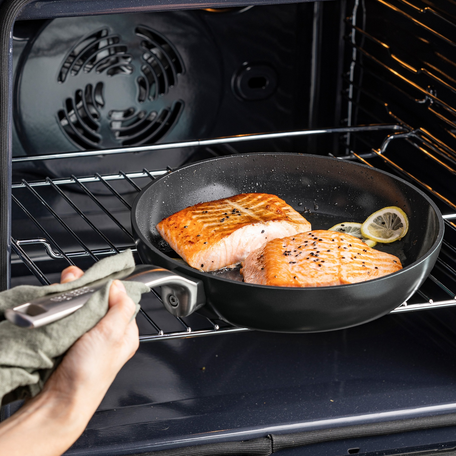 Salmon fillets in a pan inside an oven. Slices of lemon are also in the pan. A hand holds the pan handle with a cloth.