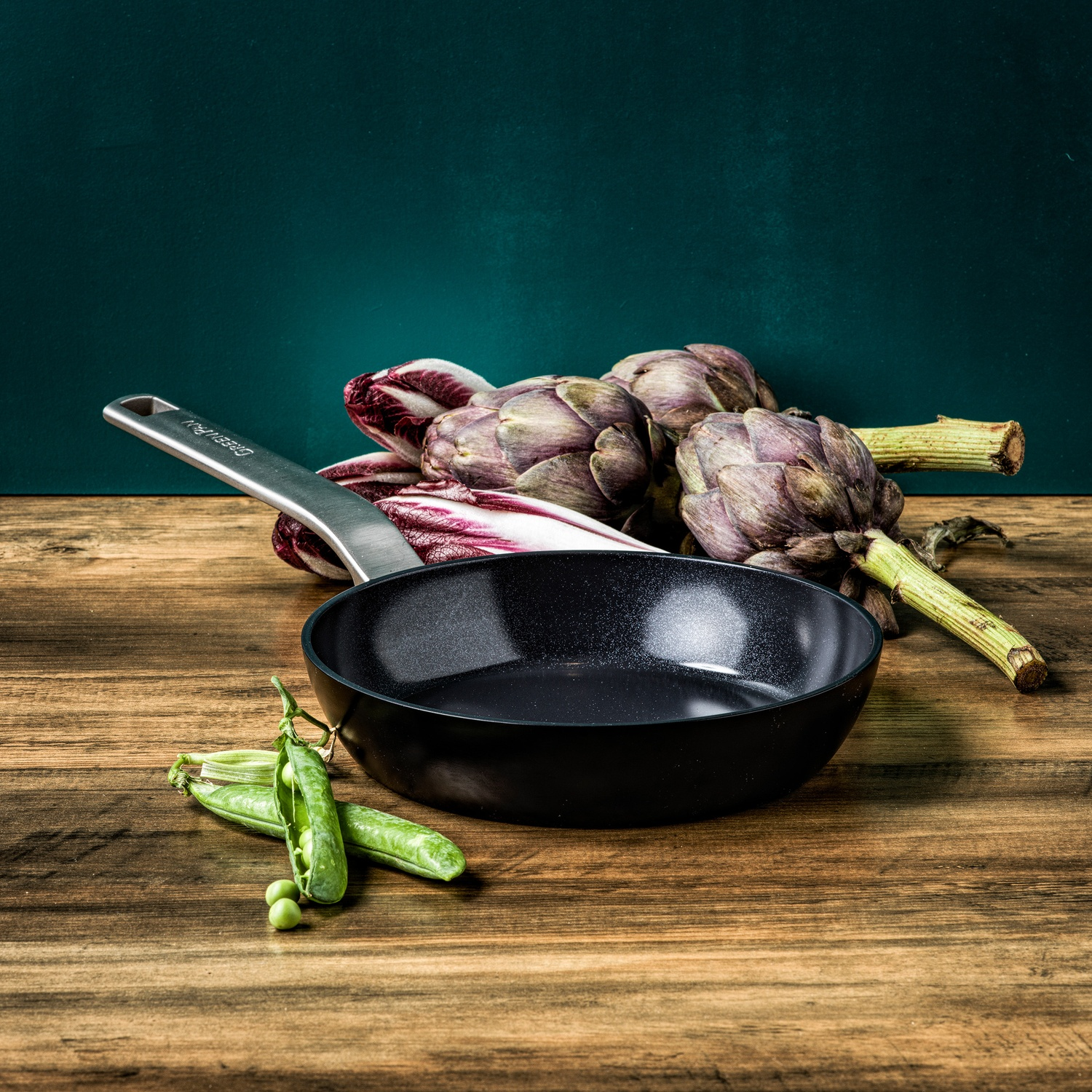 A black pan on a wooden table with artichokes, red radicchio, and pea pods. Green background.