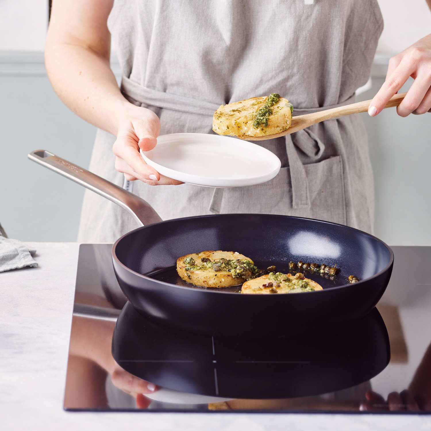 Een persoon serveert eten uit een koekenpan. Het eten is aangebraden met groene kruiden. De pan staat op een zwart fornuis.