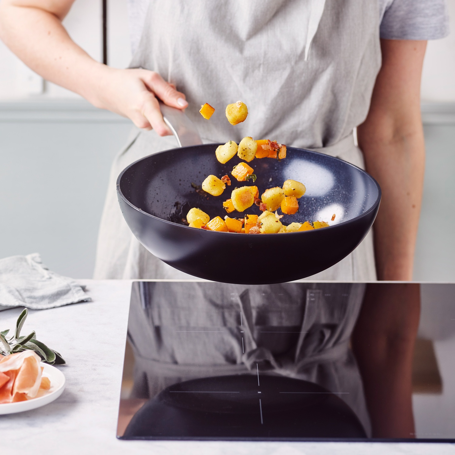 A person tosses food in a pan. The pan holds potatoes and squash. A plate of prosciutto and sage is in the background.
