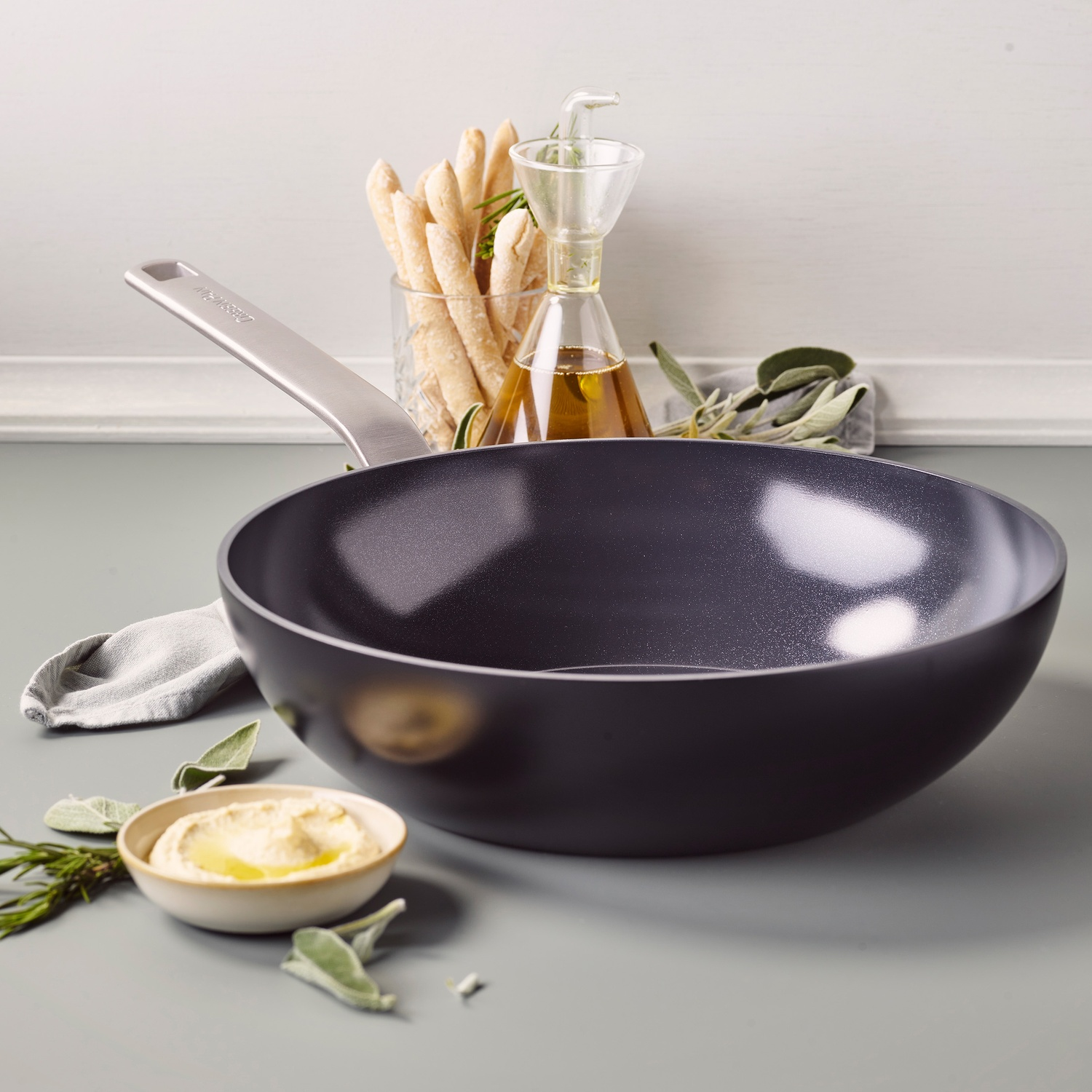 A frying pan, breadsticks, a bowl of hummus, olive oil, and herbs are arranged on a gray surface against a white backdrop.