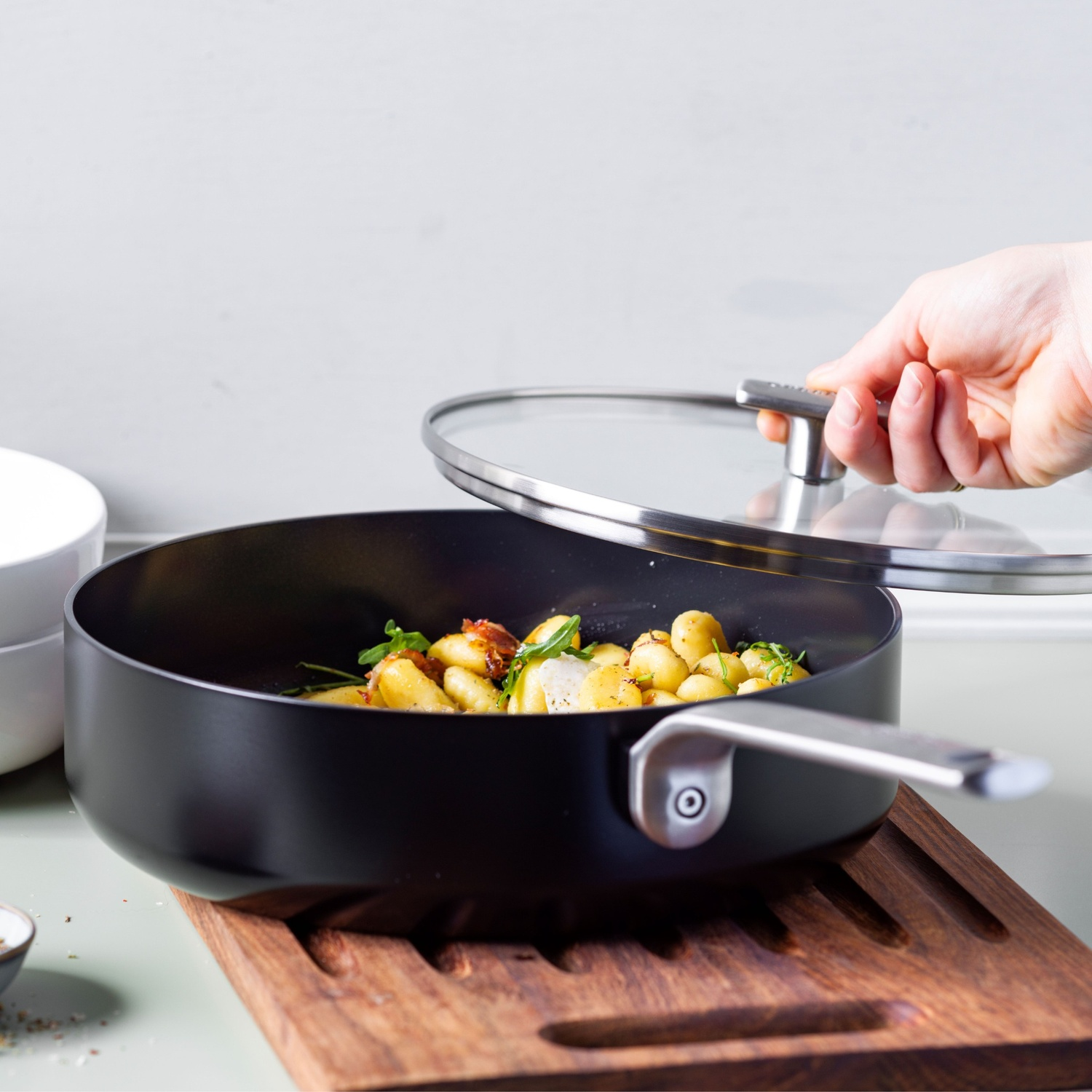 A person lifts a glass lid from a black pan filled with potatoes and herbs, placed on a wooden board.