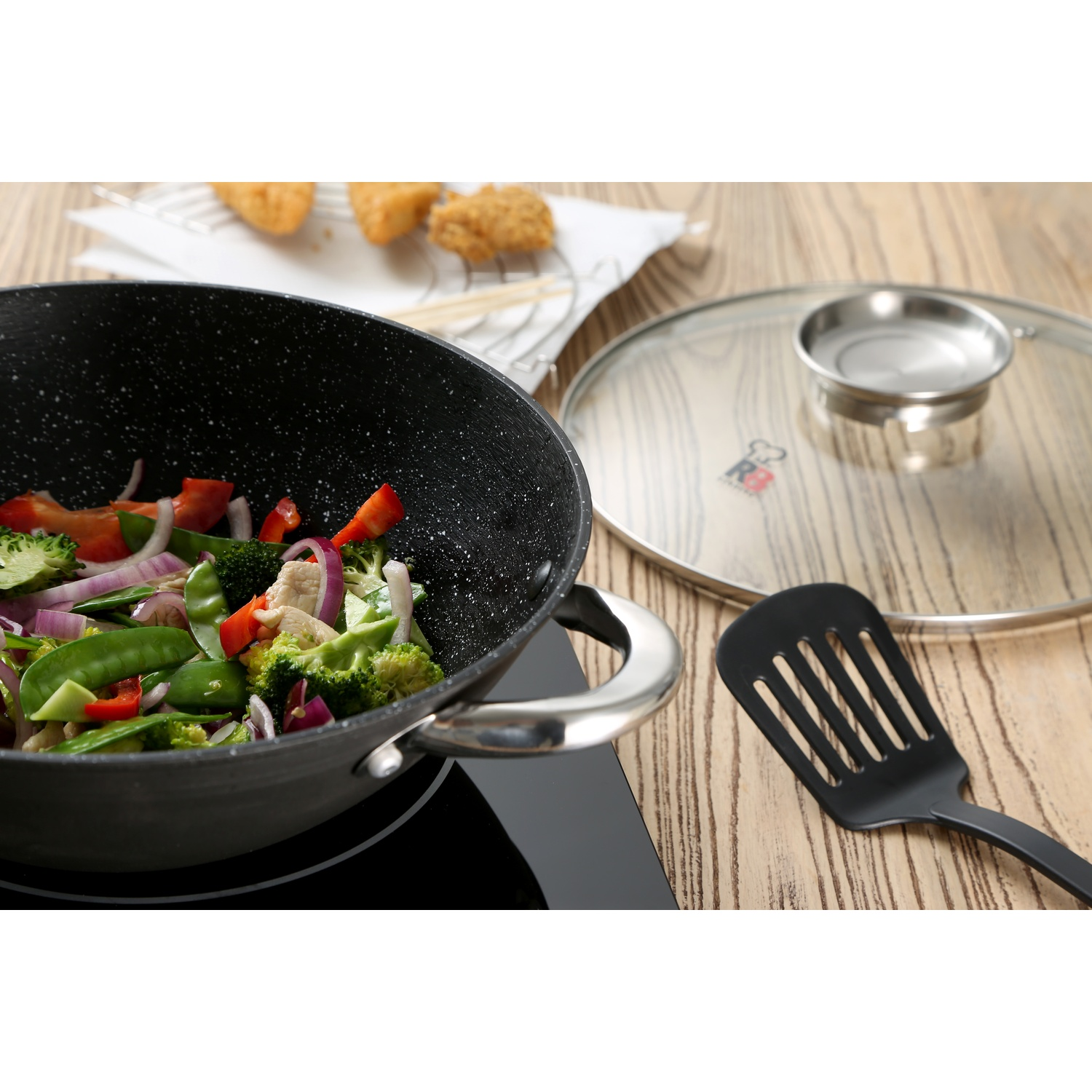 Un wok con verdure e pollo è su un piano cottura. Una spatola e un coperchio sono vicini. Cibo fritto sullo sfondo.
