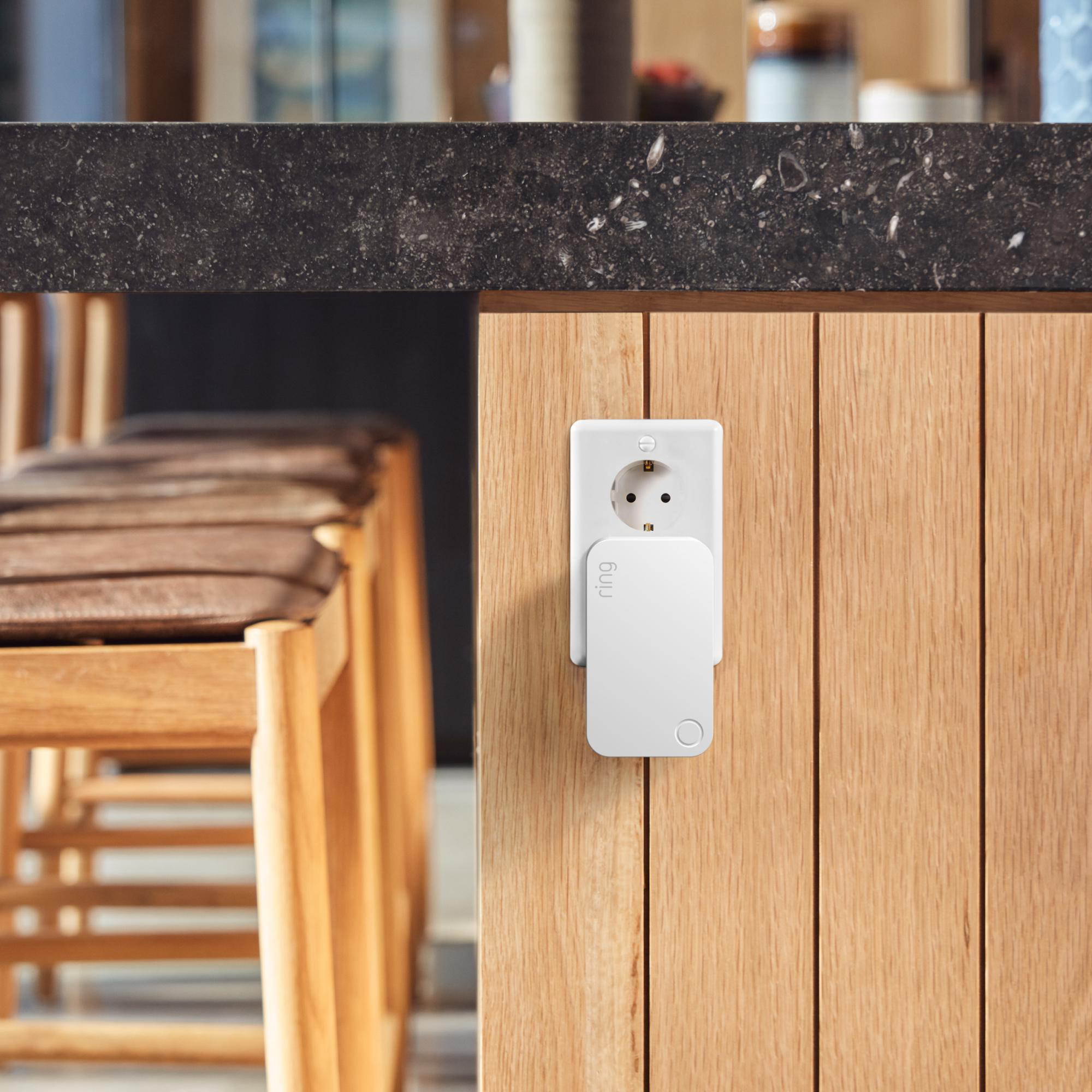 Ein weißer Ring-Smart-Stecker an einer Holz wand. Eine schwarze Arbeitsplatte und Holzstühle sind im Hintergrund.