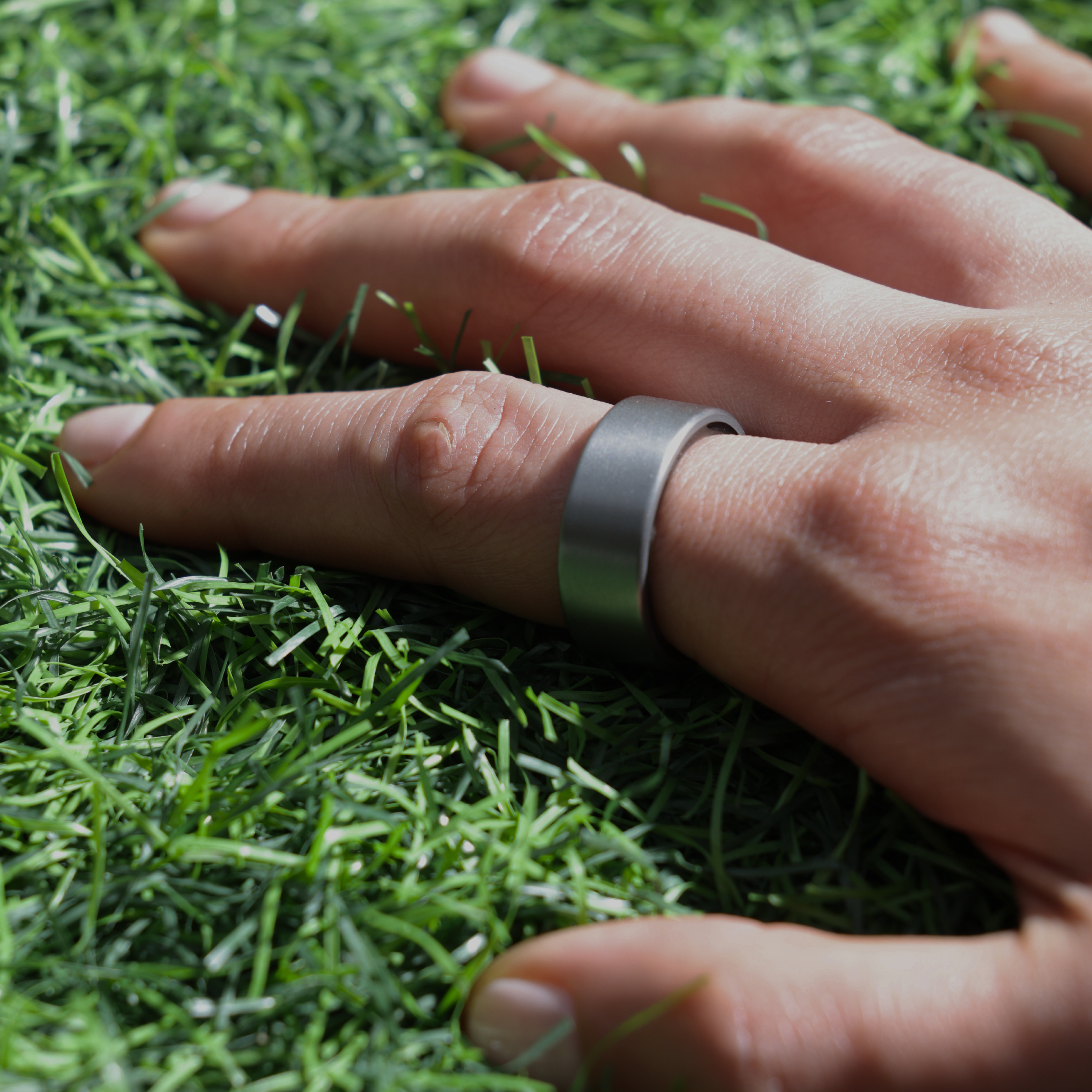 Eine Hand mit silbernem Ring auf einem Finger liegt auf grünem Kunstrasen. Heller Hautton. Weichzeichner-Hintergrund.