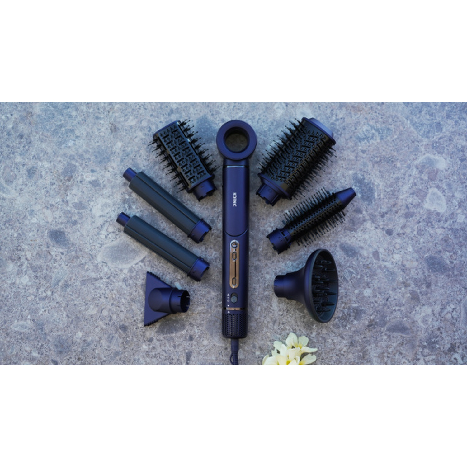 Un asciugacapelli blu scuro con accessori su una superficie di pietra. Gli accessori includono spazzole, ugelli e un diffusore. Fiori.
