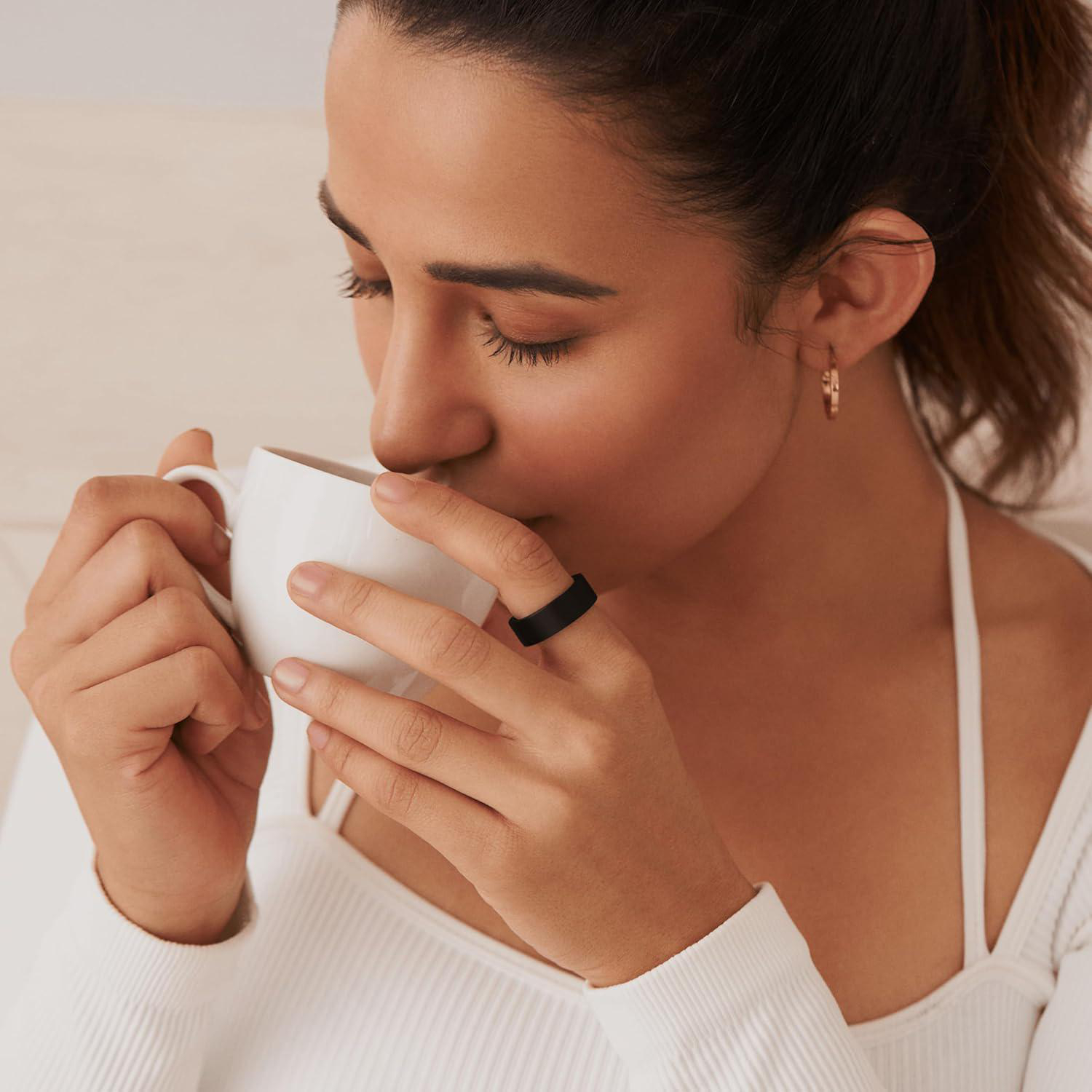 Eine Frau trinkt aus einer weißen Tasse. Sie trägt ein weißes Oberteil und einen schwarzen Ring. Ihre Augen sind geschlossen.