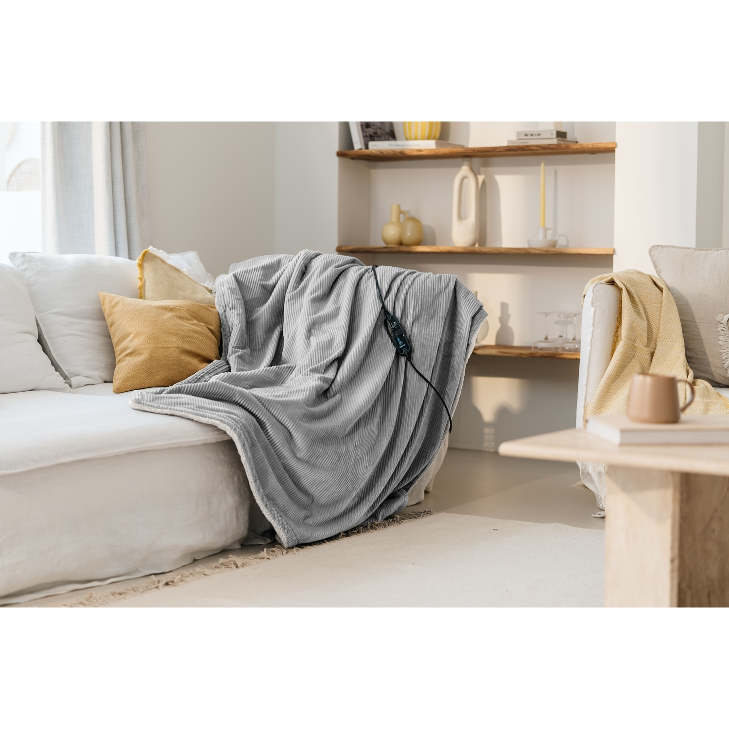 A gray blanket with corduroy texture draped over a white sofa. Yellow and white pillows are on the sofa. A wooden table and shelves are in the background.