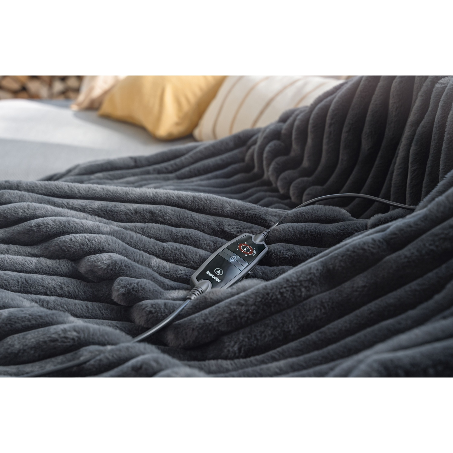 A gray, textured electric blanket with a control panel is placed on a bed. Pillows and firewood are visible in the background.